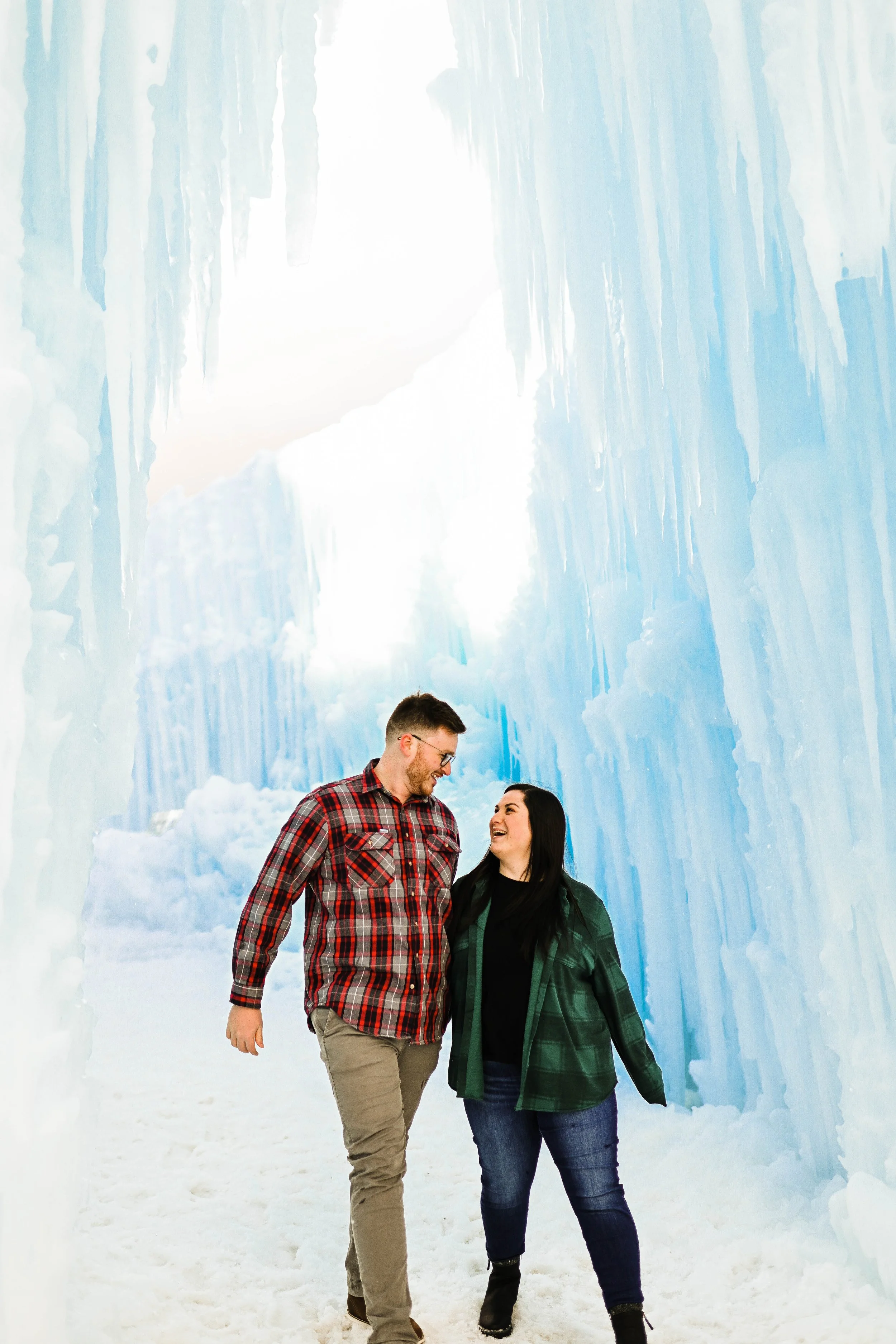 A man and woman smiling and walking together inside an ice cave with walls made of blue ice and snow on the ground.