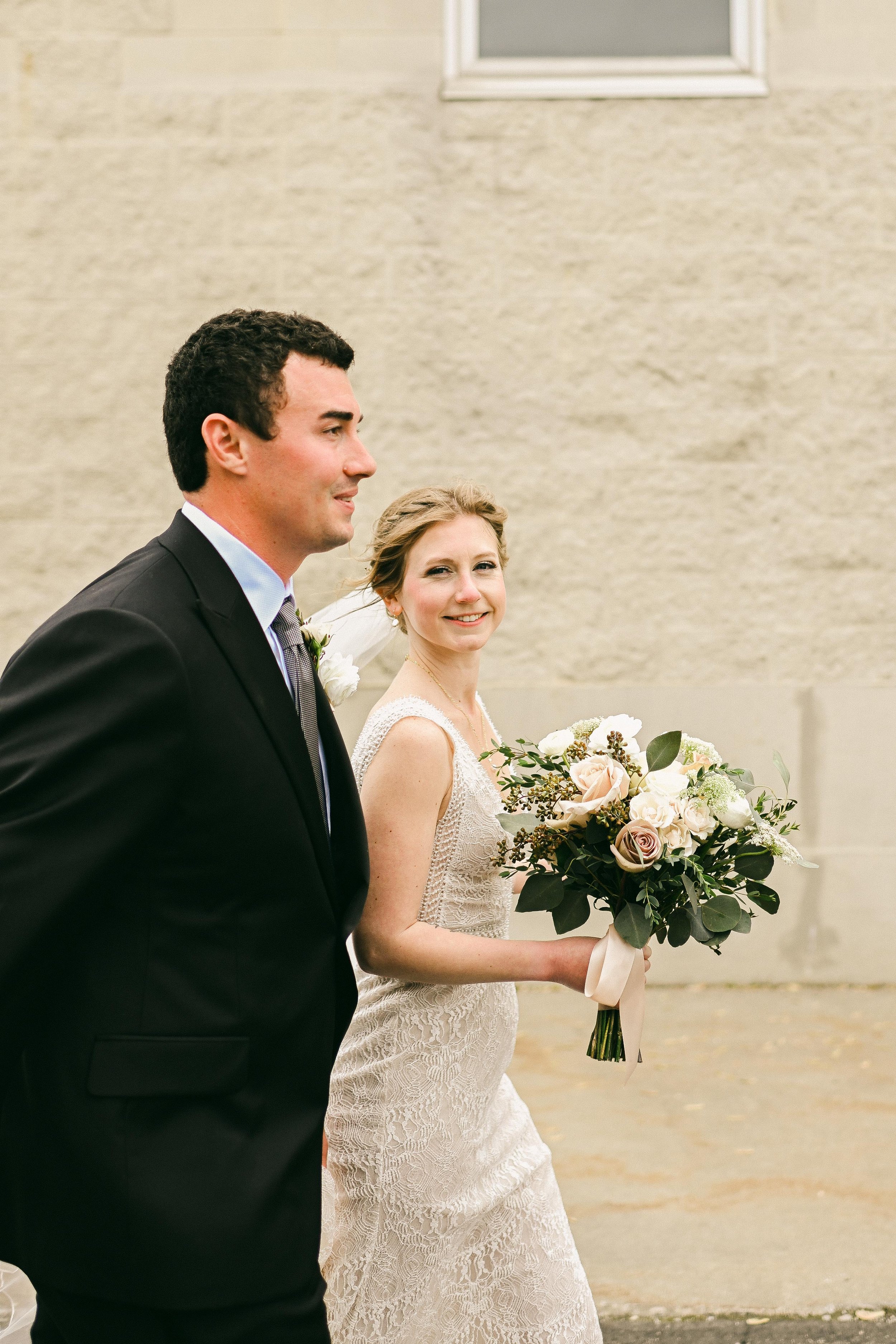 A bride in a lace wedding dress holding a bouquet of white and blush roses, walking beside a groom in a black suit, outside near a beige wall.
