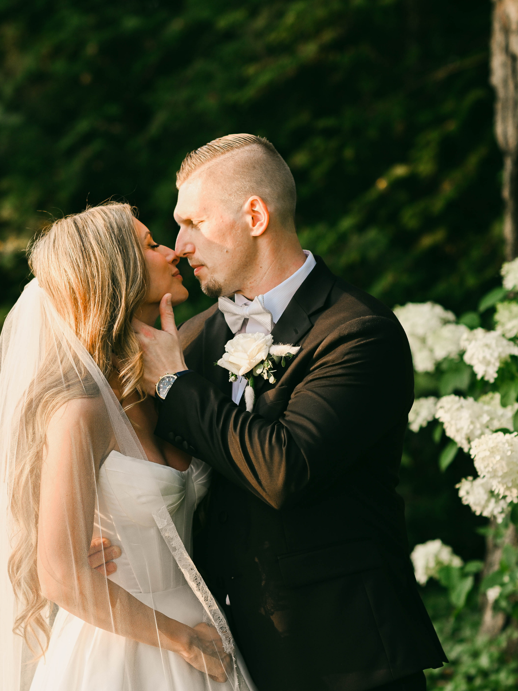 Bride and groom in an intimate moment during their wedding, standing outdoors with greenery and white flowers in the background.