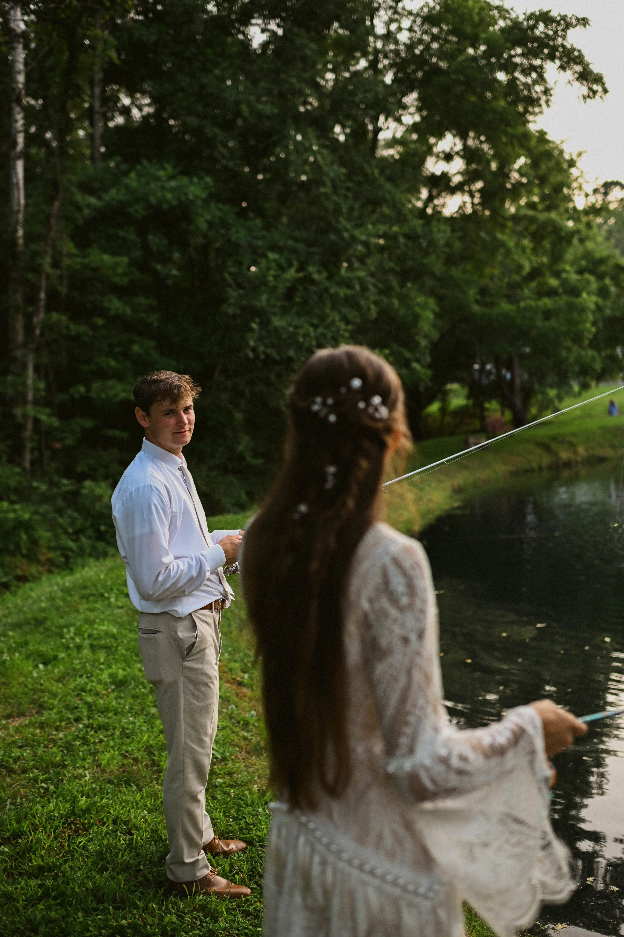 A young man in a white shirt and beige pants standing by a river, and a woman with long brown hair in a white lace dress with floral beads in her hair, fishing by the river in a park surrounded by tall trees.