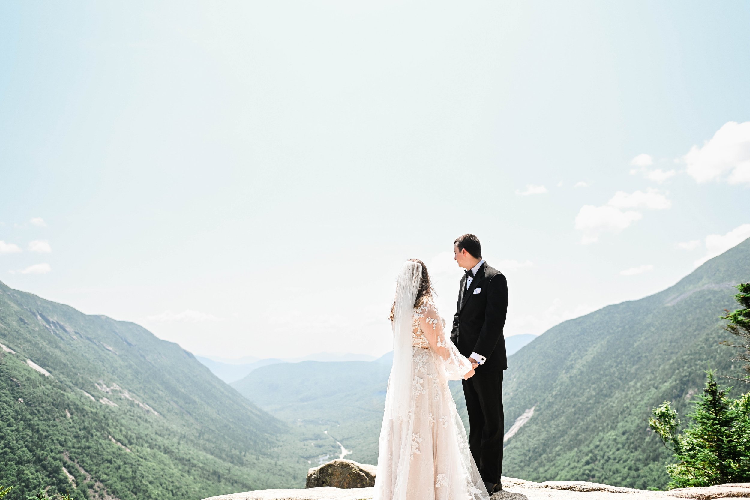 A bride and groom holding hands and facing each other outdoors on a mountain overlook, with green mountains and a bright sky in the background.