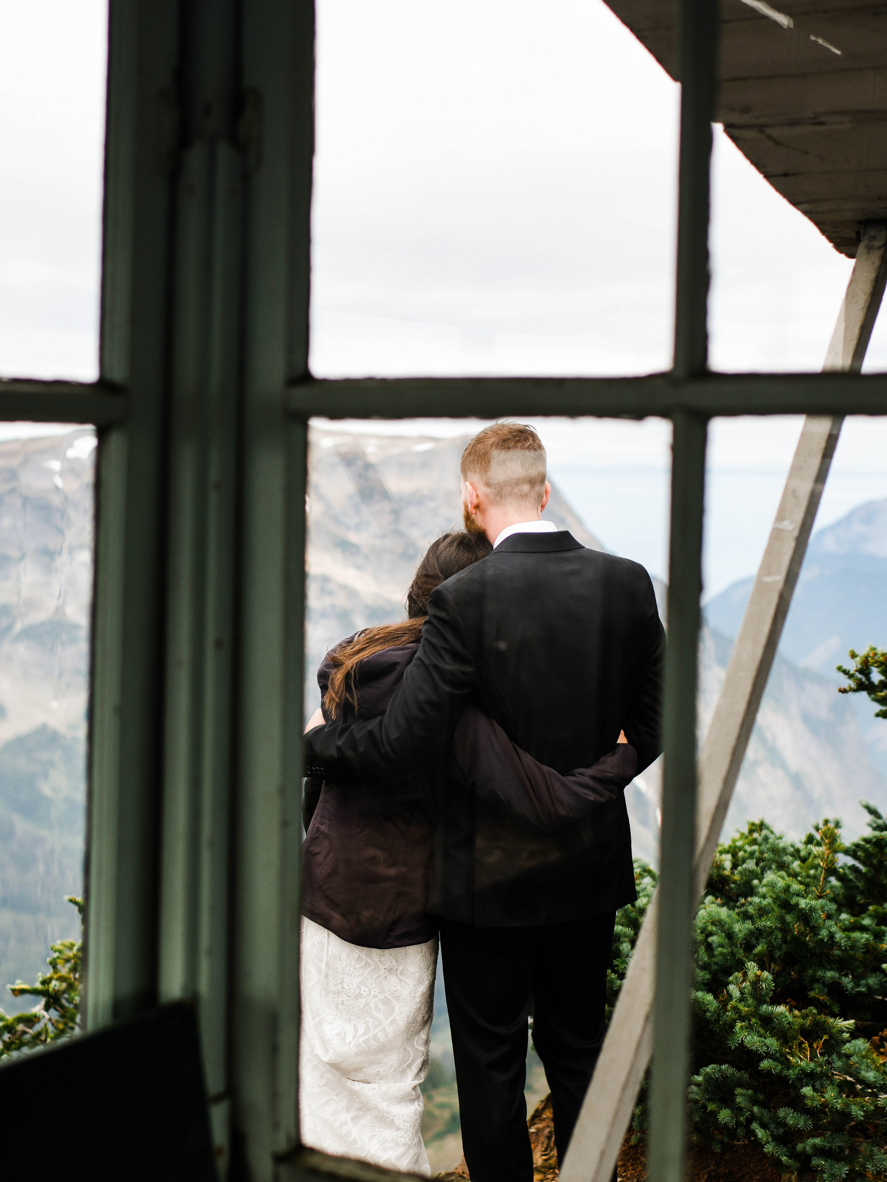 A couple embracing outdoors, viewed through a window frame, with mountains and greenery in the background.