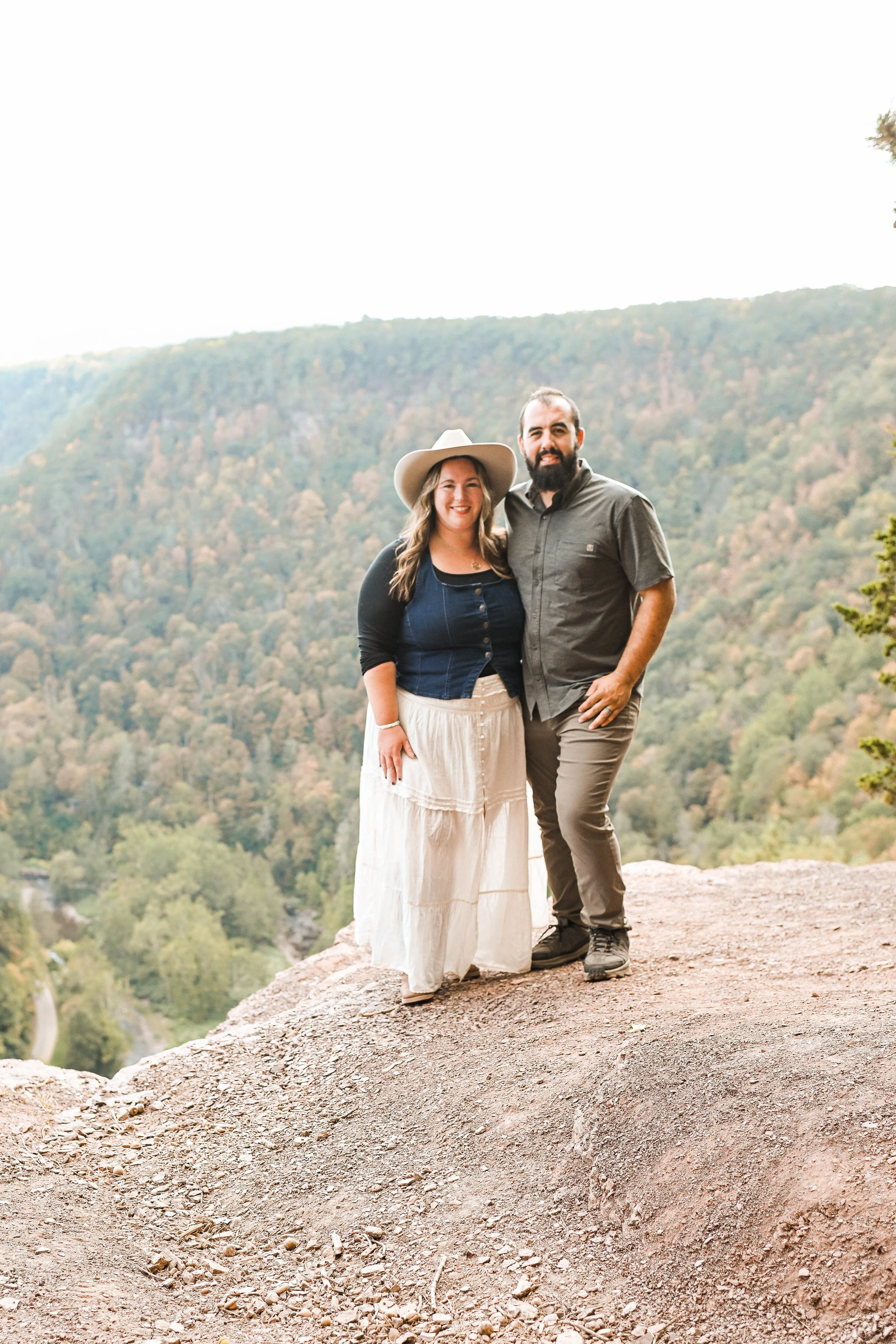 A couple standing on a rocky ledge outdoors with a forested valley in the background. The woman is wearing a wide-brimmed hat, a dark blue top, and a long white skirt. The man is dressed in a gray button-up shirt and khaki pants. They are smiling at the camera.