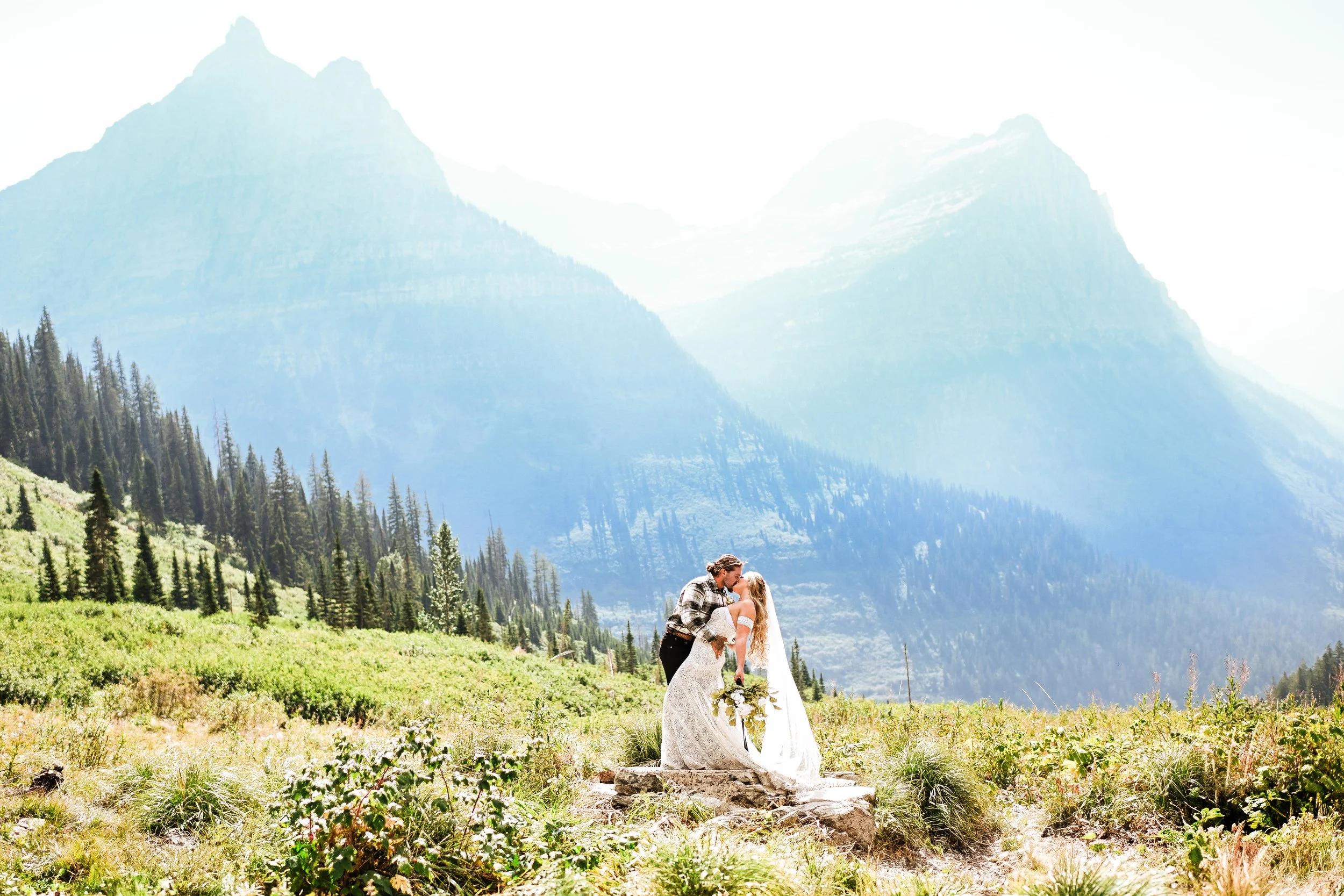 A couple dressed in wedding attire sharing a kiss outdoors in a mountainous landscape with tall trees and green fields.