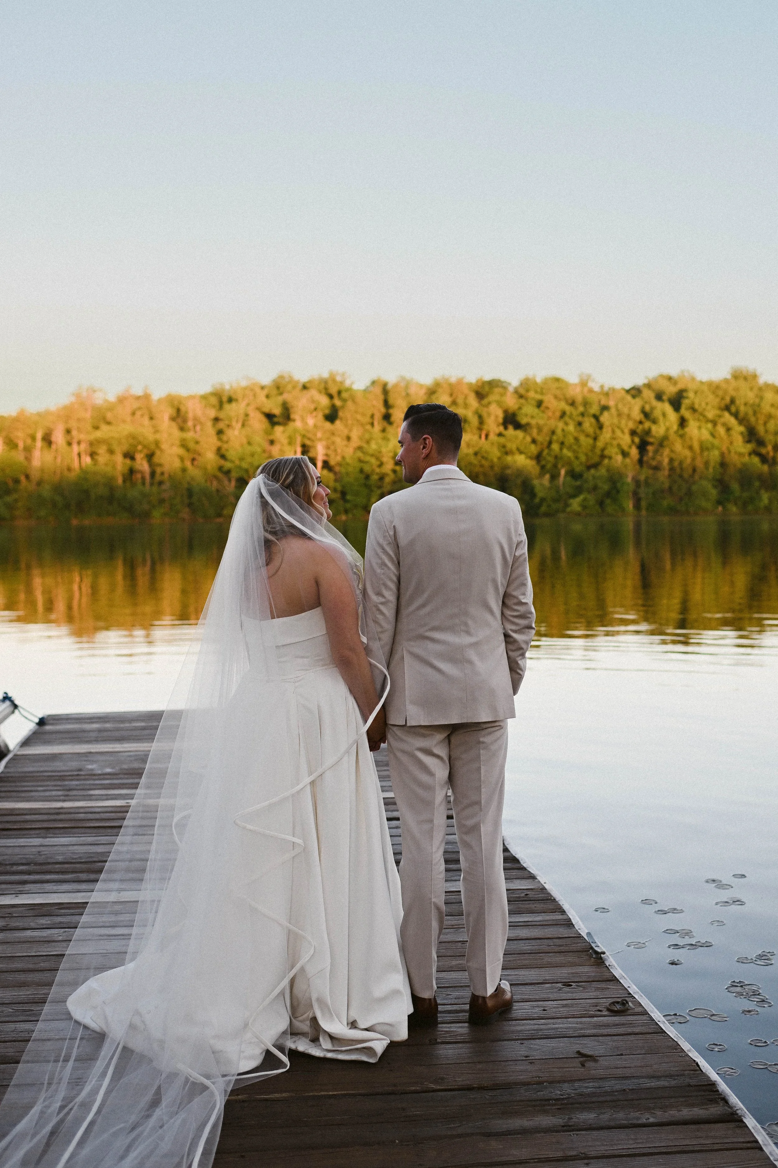 A bride and groom standing hand in hand on a wooden dock by a lake at sunset, with trees in the background, facing each other and smiling.