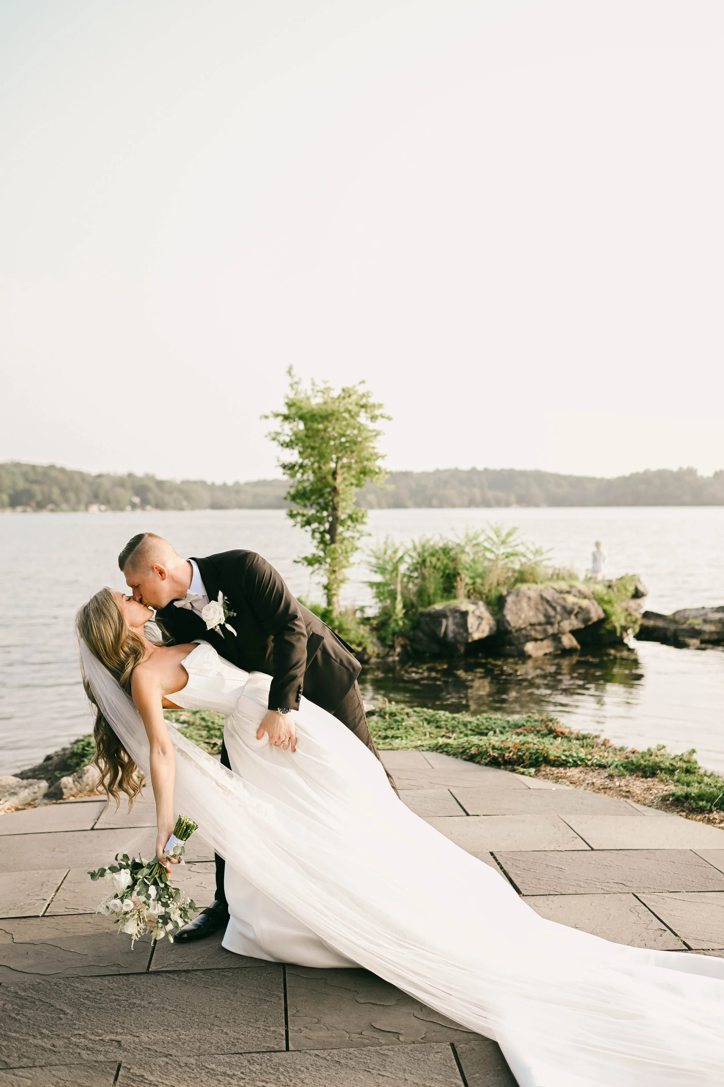 A newlywed couple sharing a kiss by a lakeside, with the groom leaning over the bride who is holding a bouquet, on a paved area with water and rocks in the background.