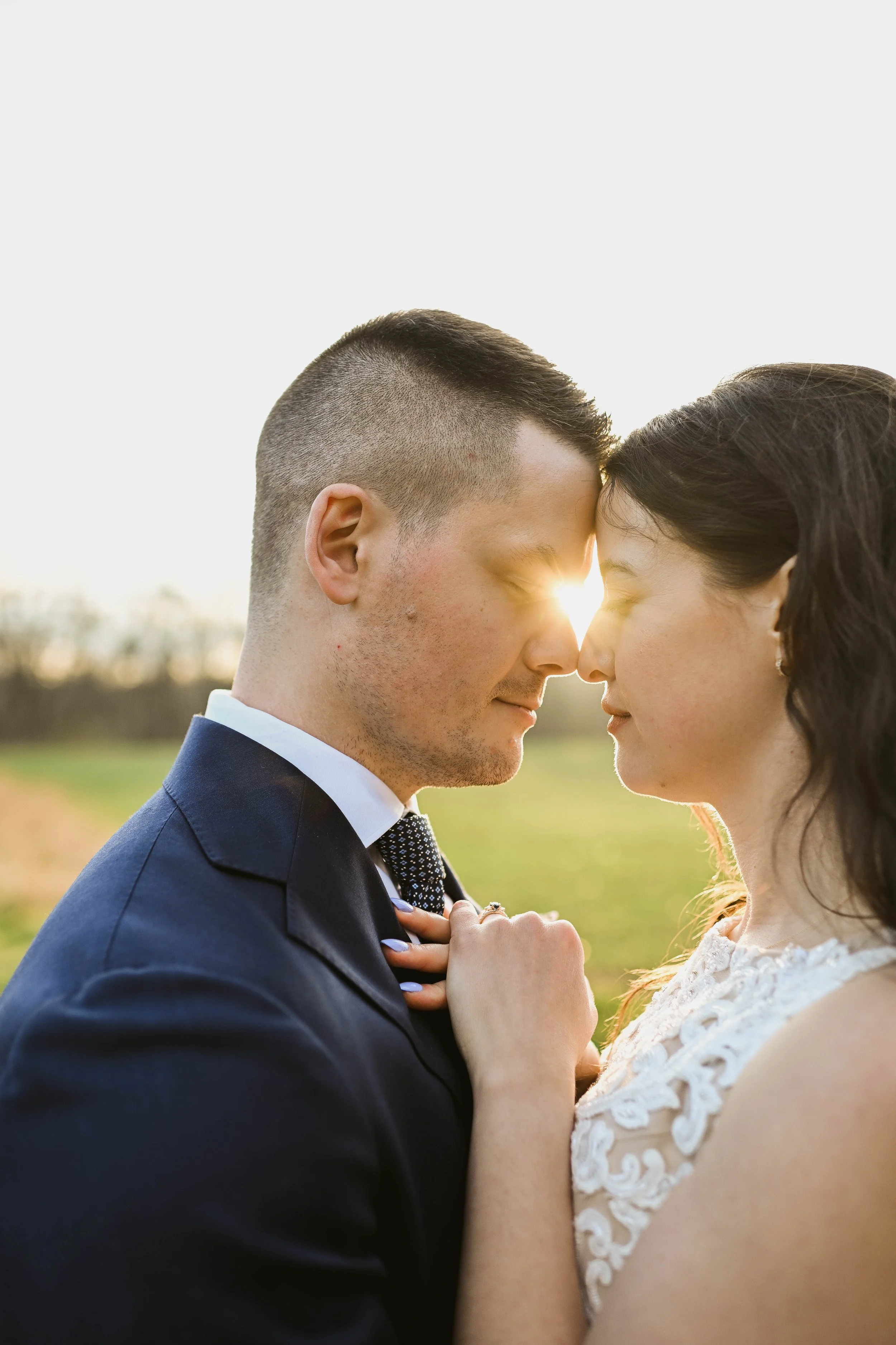 A couple, with their foreheads touching, standing outdoors in a field during sunset, with the sun between their faces, creating a warm glow.