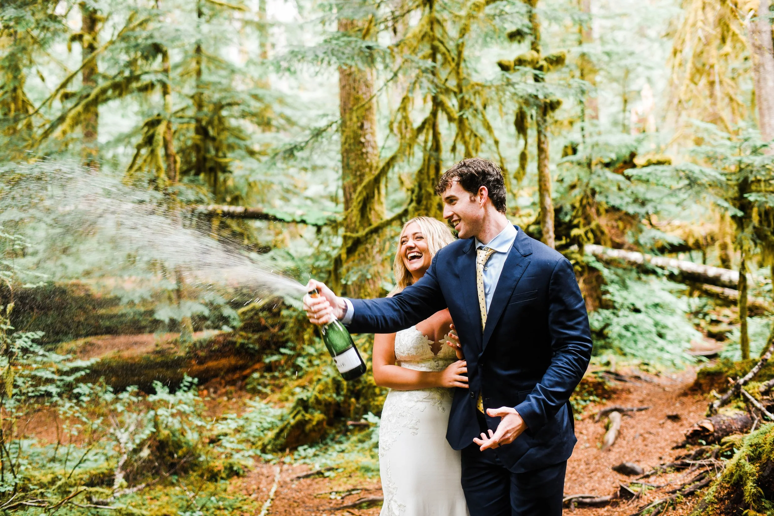 A couple celebrating in a forest, with the man in a suit opening a champagne bottle, causing it to spray, and the woman in a white dress smiling.