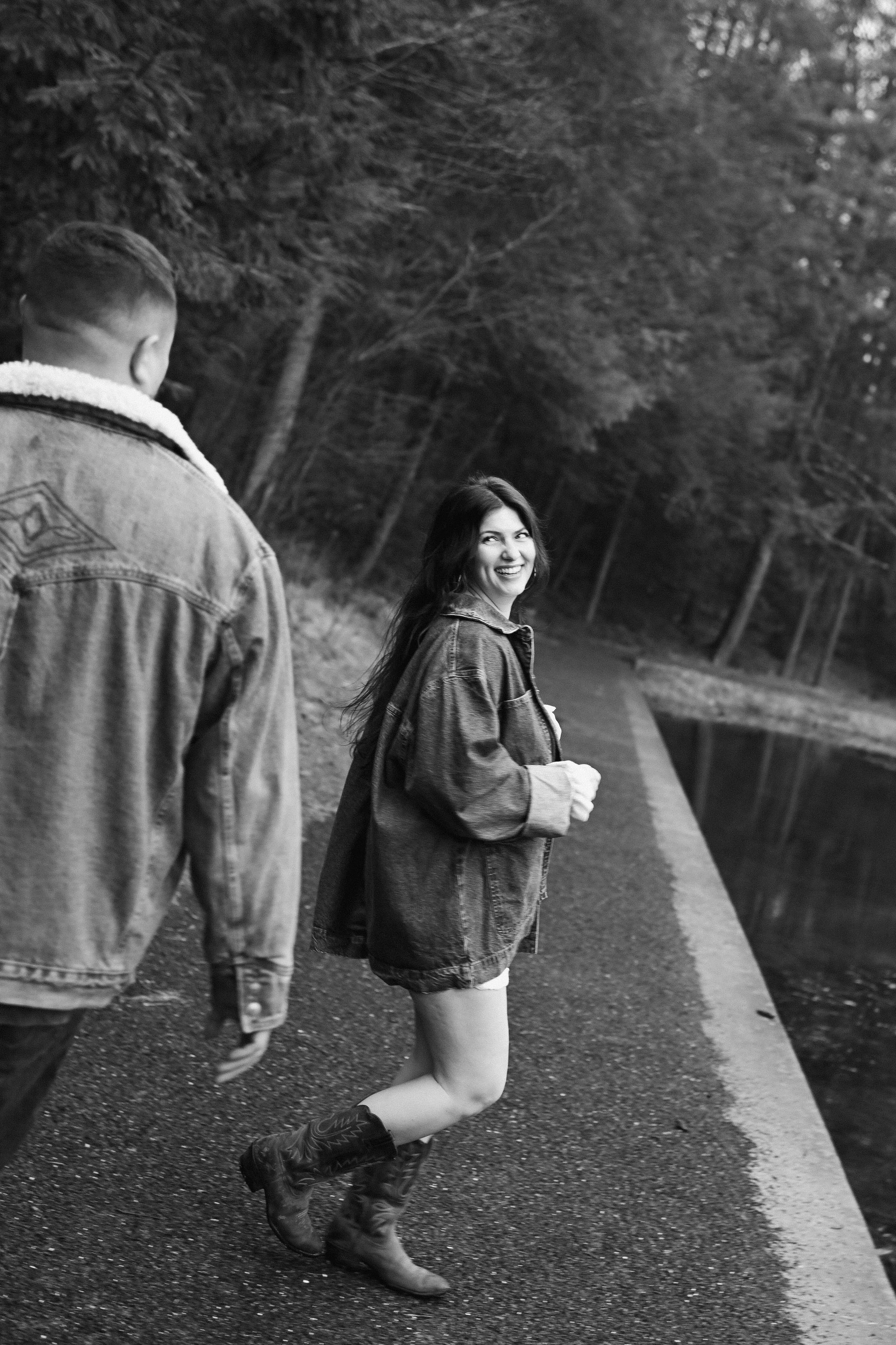 A woman smiling and walking on a paved path beside a body of water, with a man walking alongside her, surrounded by trees.