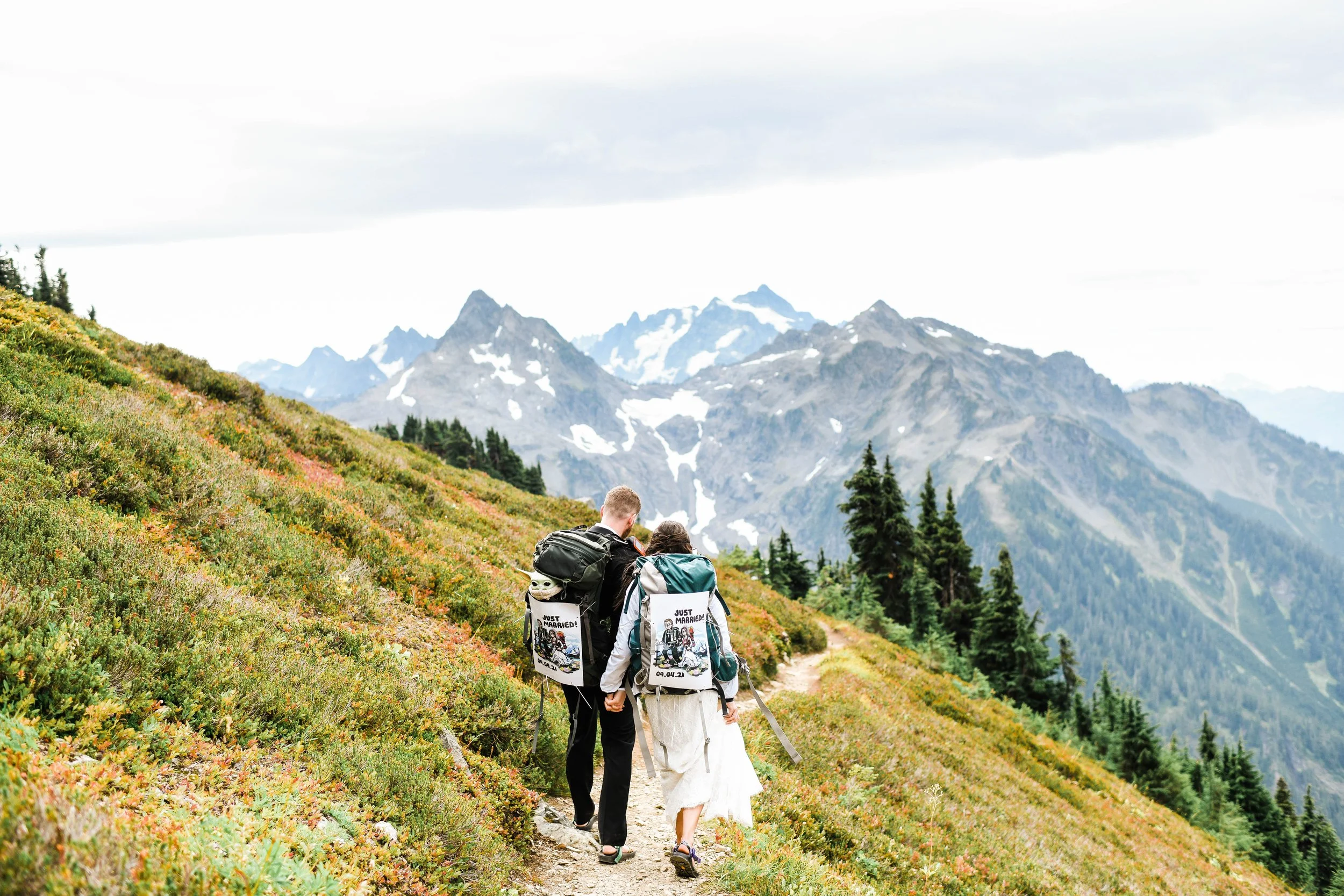 A couple hiking on a mountain trail with snow-capped peaks in the background, surrounded by green and colorful vegetation.