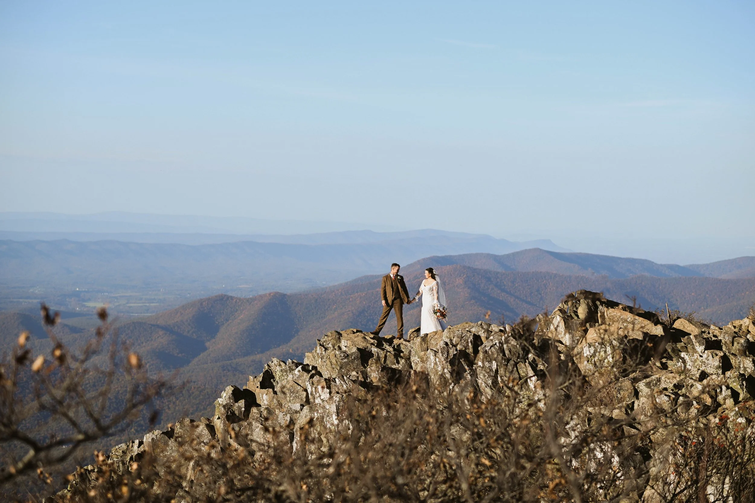 A bride and groom holding hands on a rocky mountain ridge, with a scenic view of distant mountains and a clear blue sky in the background.