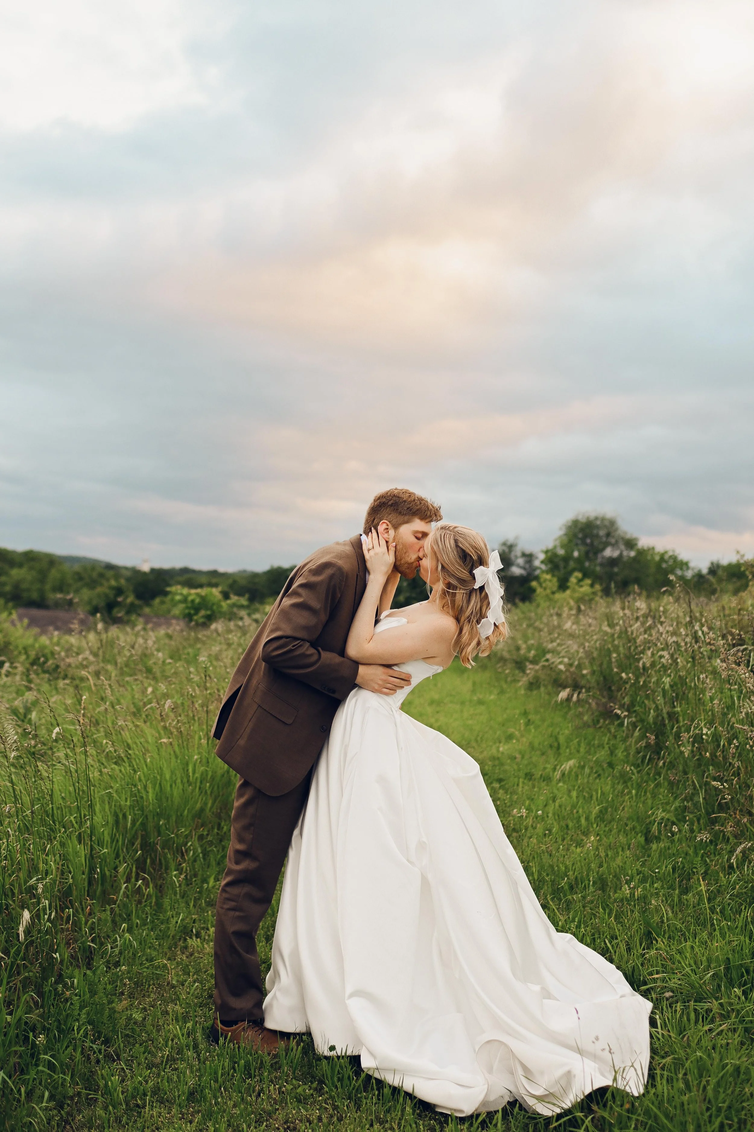 A newlywed couple kissing in a grassy field during sunset, the bride wearing a white wedding gown with a bow in her hair, and the groom in a brown suit.