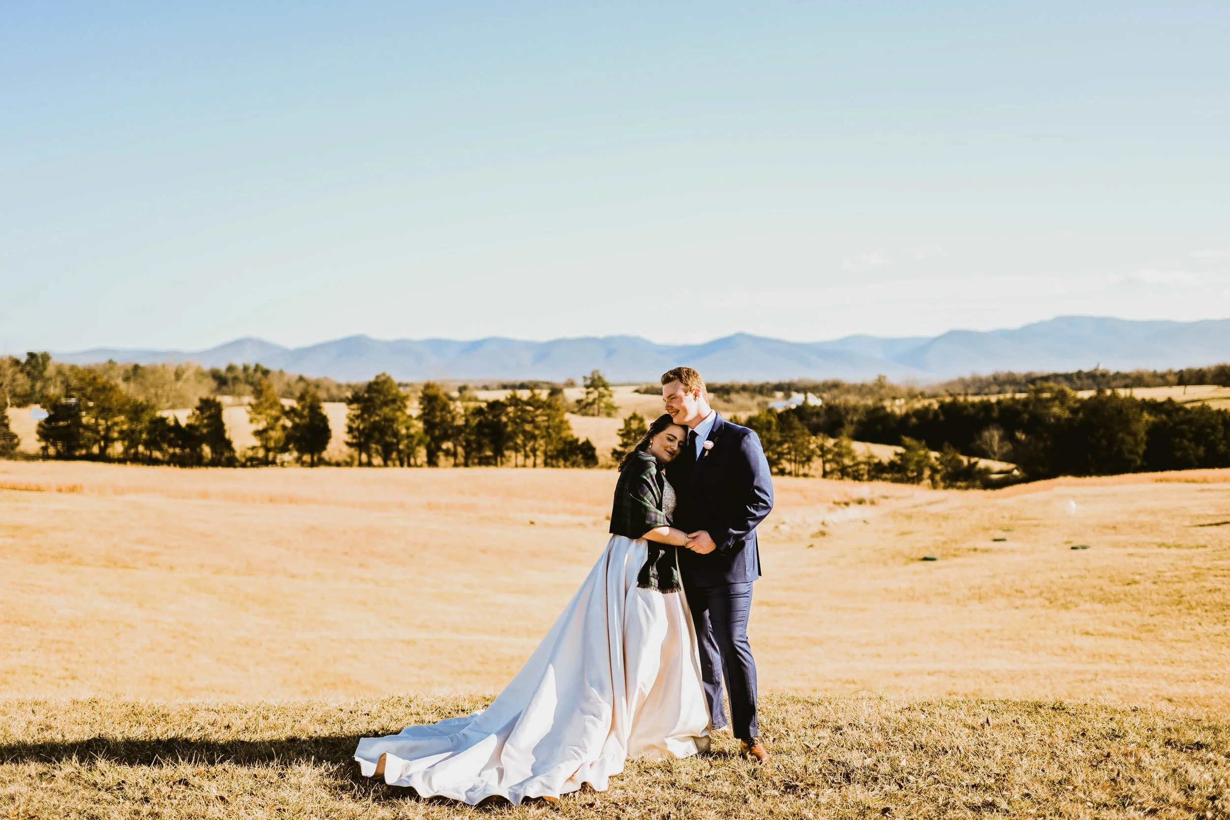A couple dressed in wedding attire standing in a field with a scenic landscape of trees and mountains in the background.
