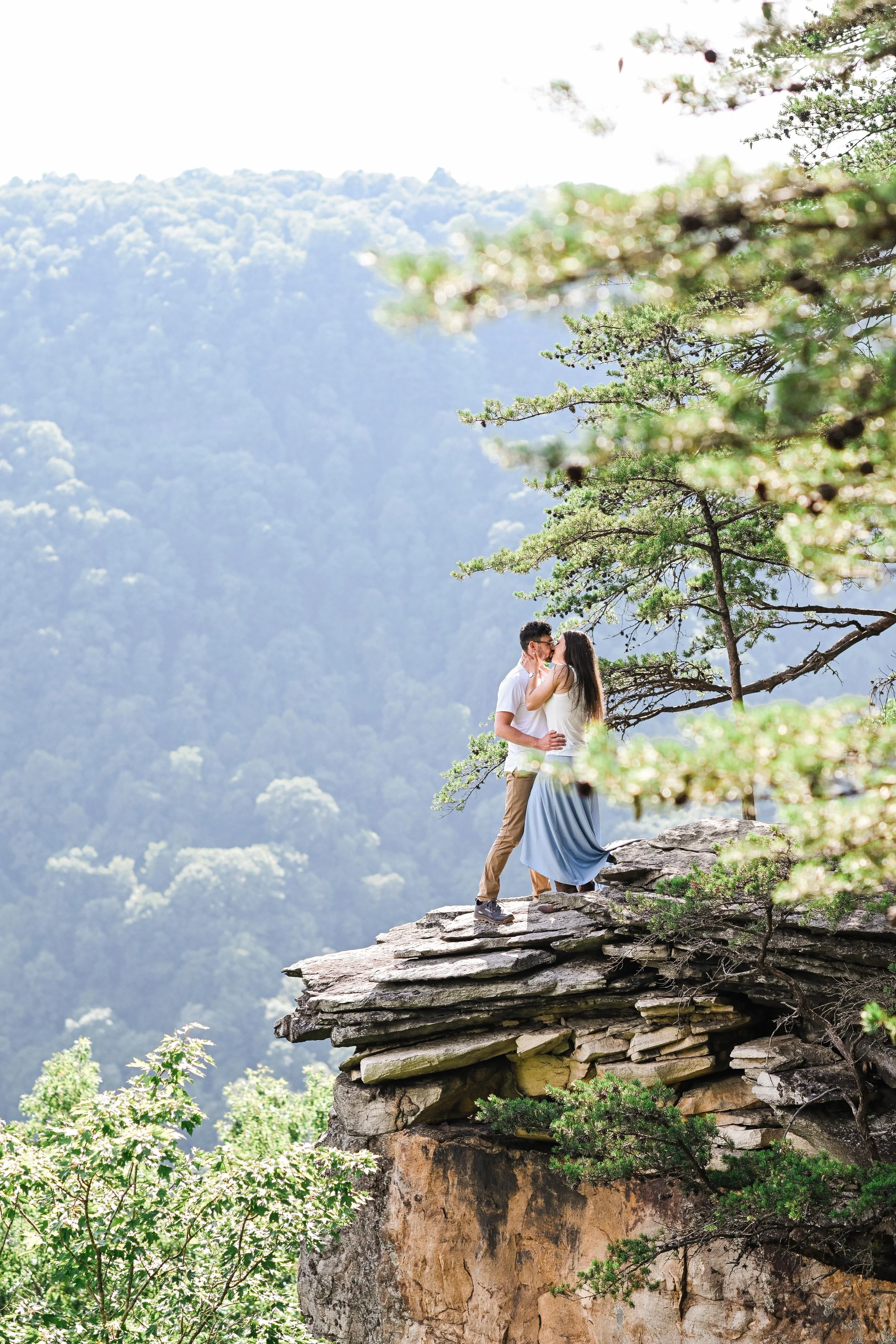 A couple kissing on a rocky ledge with green trees and mountain background.