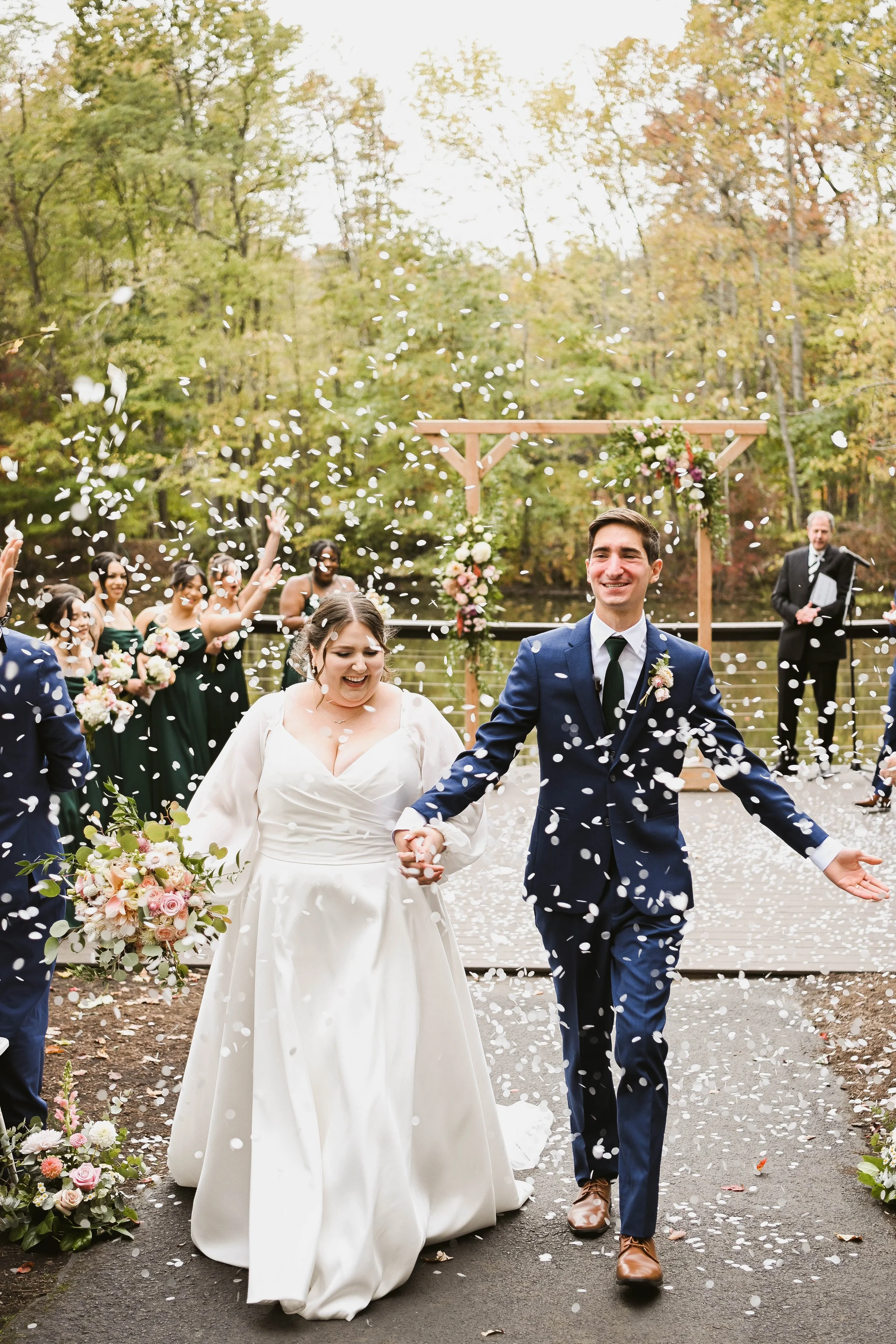 A newlywed couple walking hand in hand through a celebration, surrounded by friends and family, with falling flower petals, outdoors in a wooded area during daytime.
