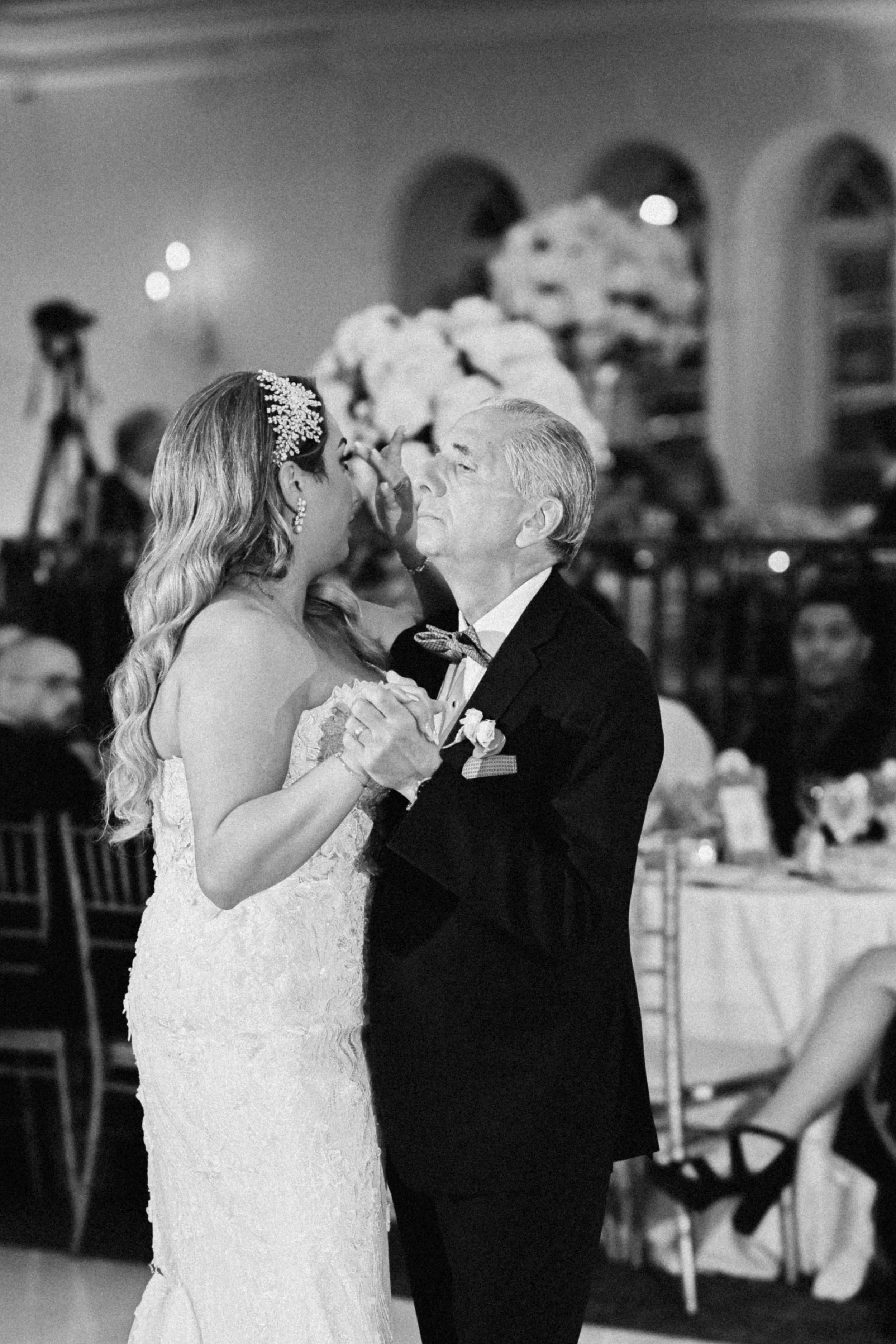 A bride dancing with an elderly man at her wedding reception. The bride is wearing a strapless wedding dress with a floral headpiece, and the man is in a tuxedo. They are holding hands and dancing closely, with a floral centerpiece and decorated tabl