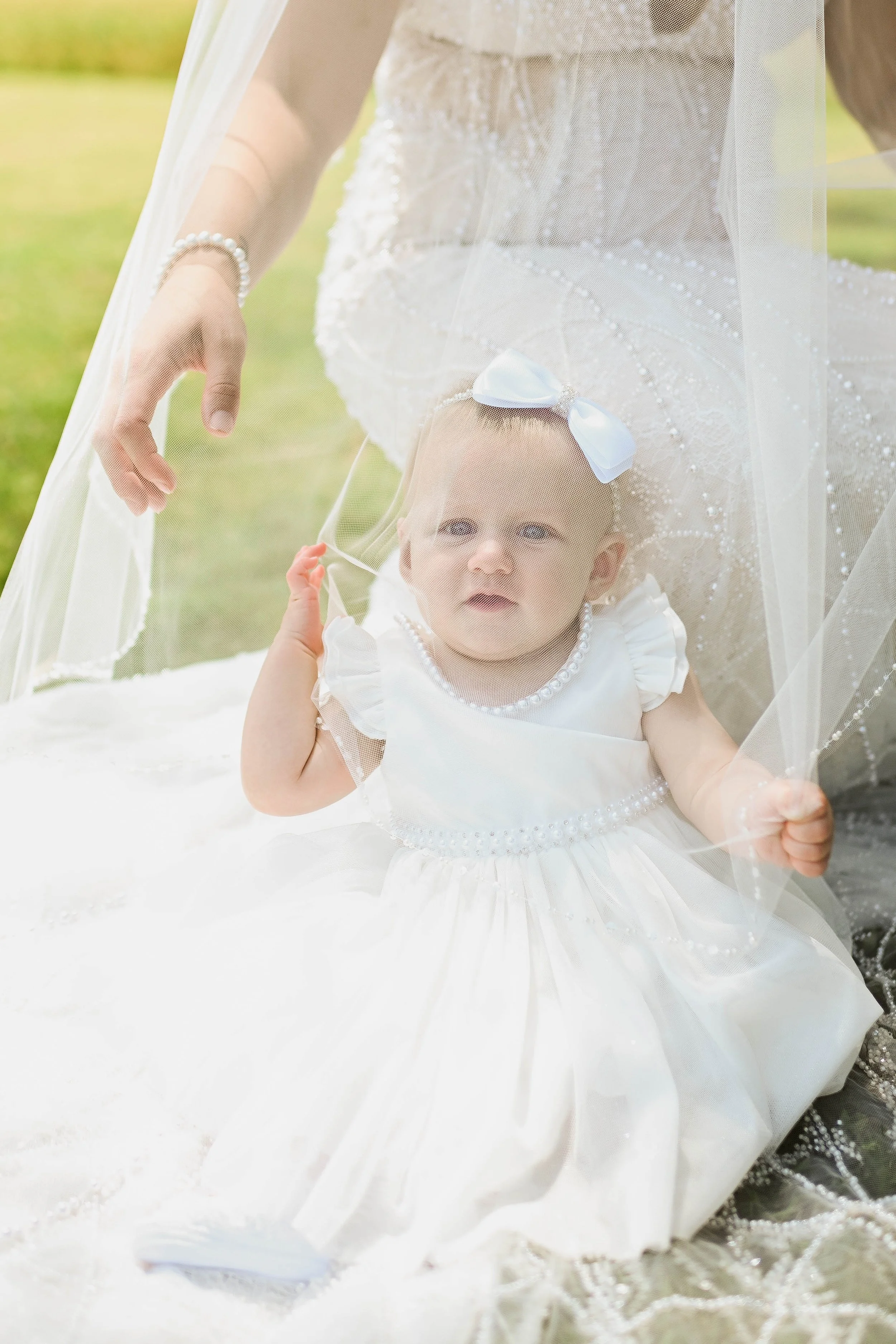 Close-up of a baby girl in a white dress with pearl details, sitting on a white surface. She has a white bow headband, and is partly covered by a veil. An adult's hand is adjusting the veil. The background is outdoor with greenery.