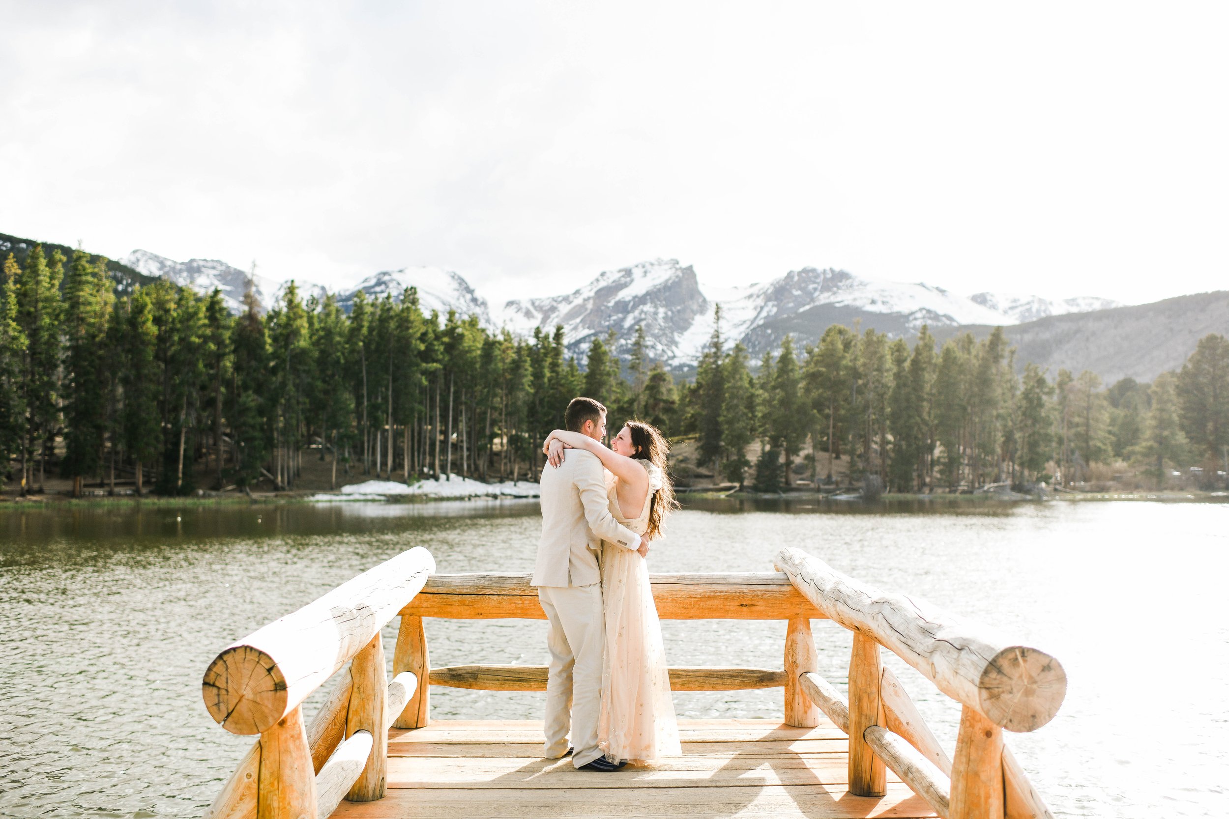 A couple stands on a wooden dock embracing, with a mountain lake and snow-capped mountains in the background.