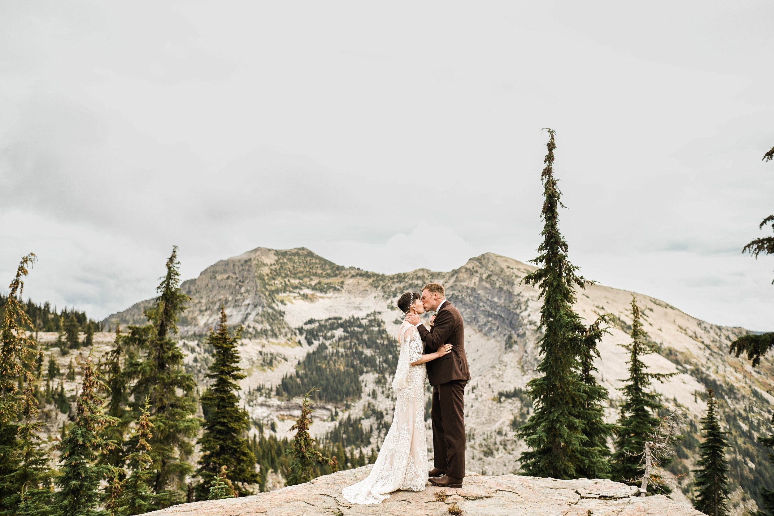 A bride and groom kissing on a rocky ledge outdoors with mountainous landscape and pine trees in the background.