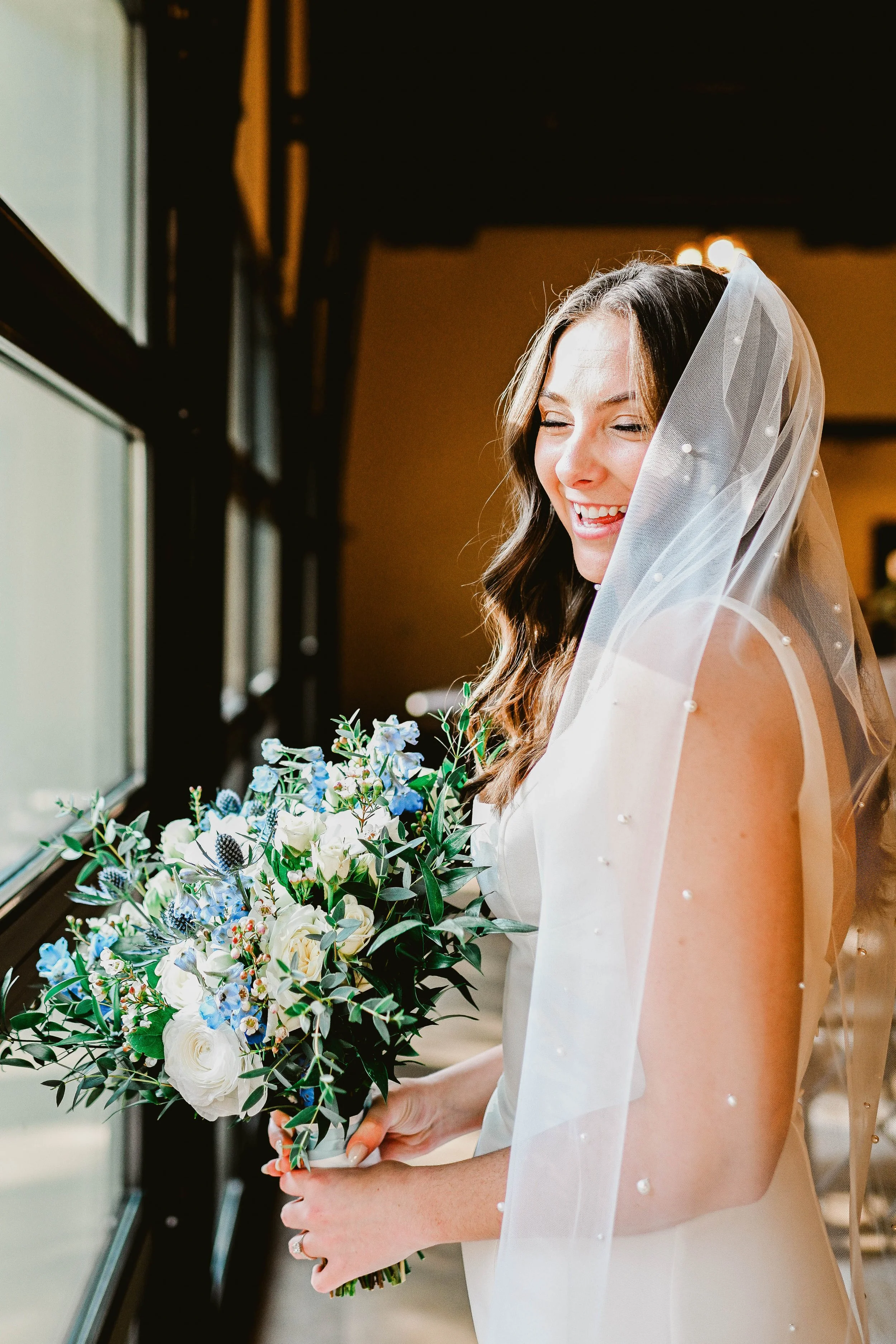 A bride with long brown hair, wearing a white wedding dress and a sheer veil with pearls, holding a bouquet of white and blue flowers, smiling and looking out of a window.