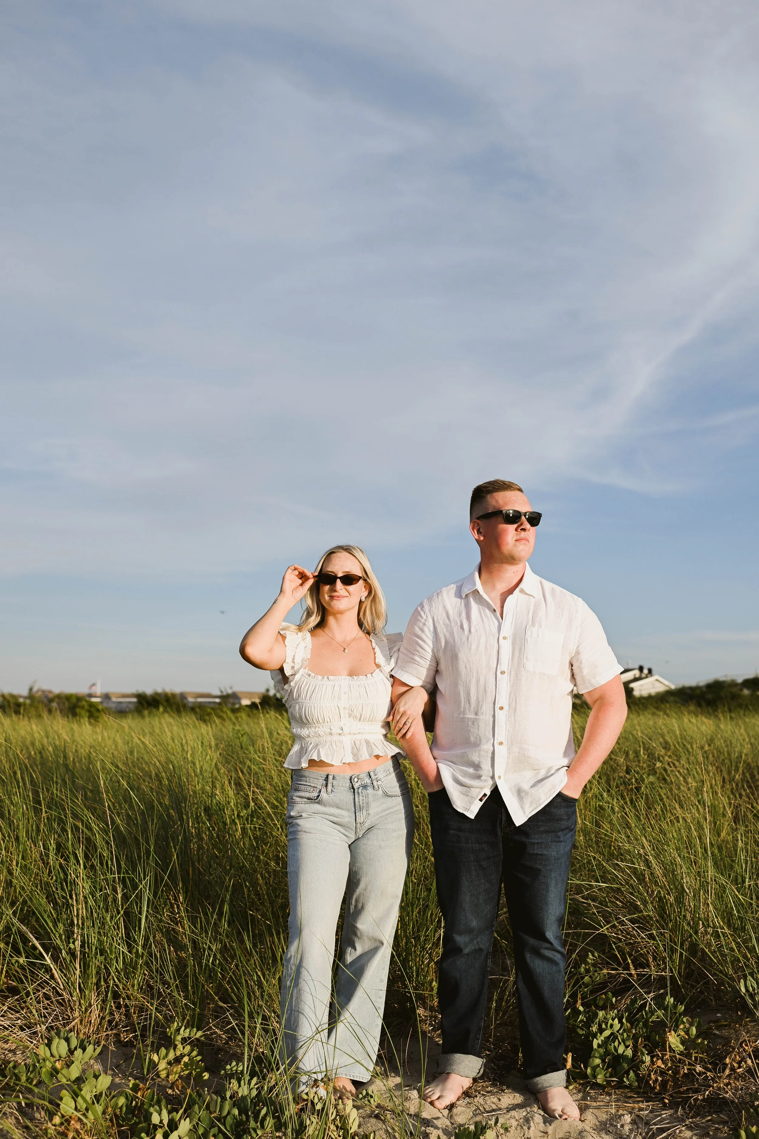 A man and woman stand hand-in-hand in a grassy field under a blue sky with wispy clouds, both wearing sunglasses and casual summer clothing.