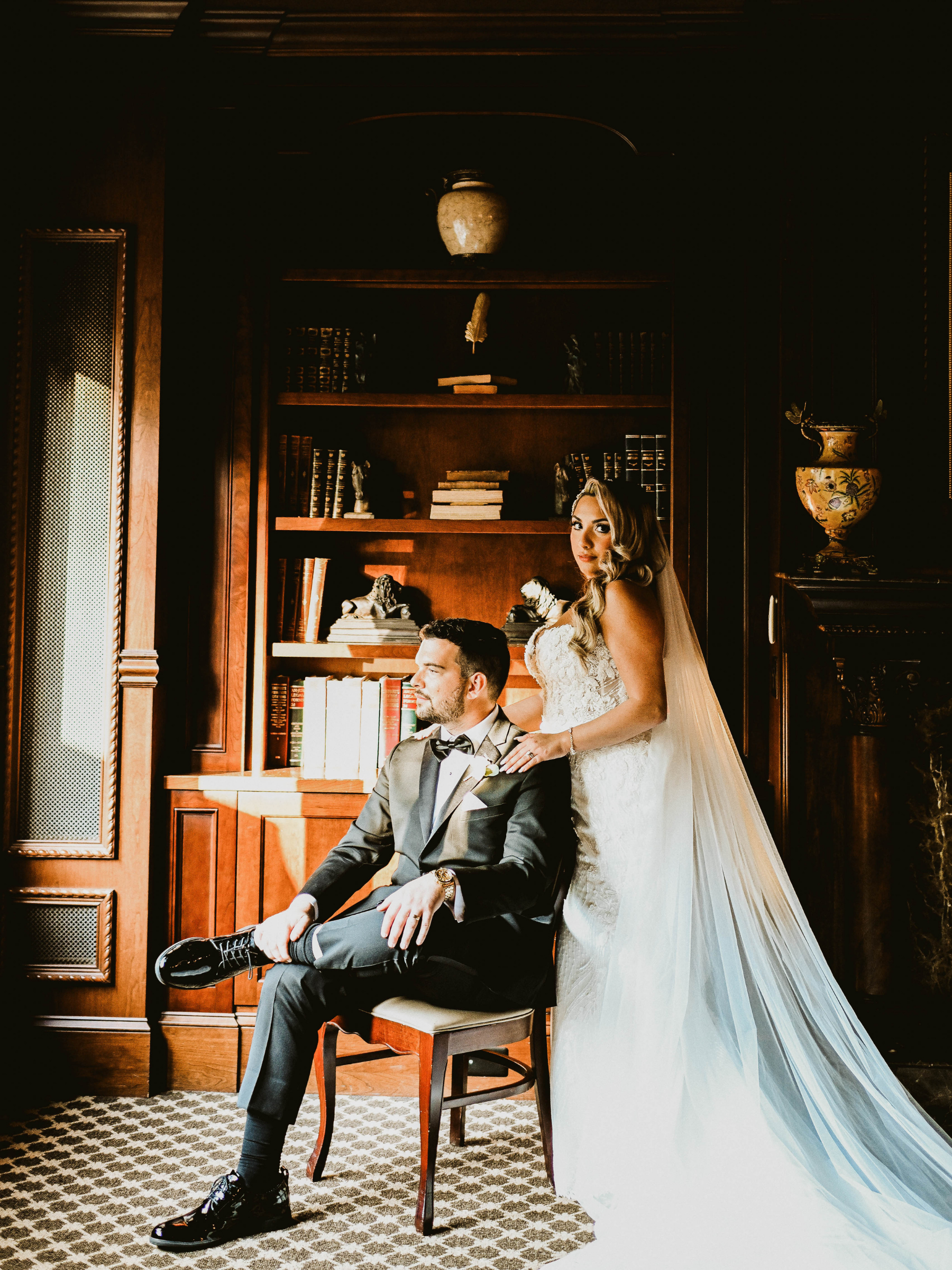 A bride in a white wedding dress with a long veil standing behind a groom in a tuxedo sitting on a chair in a wood-paneled room with bookshelves.