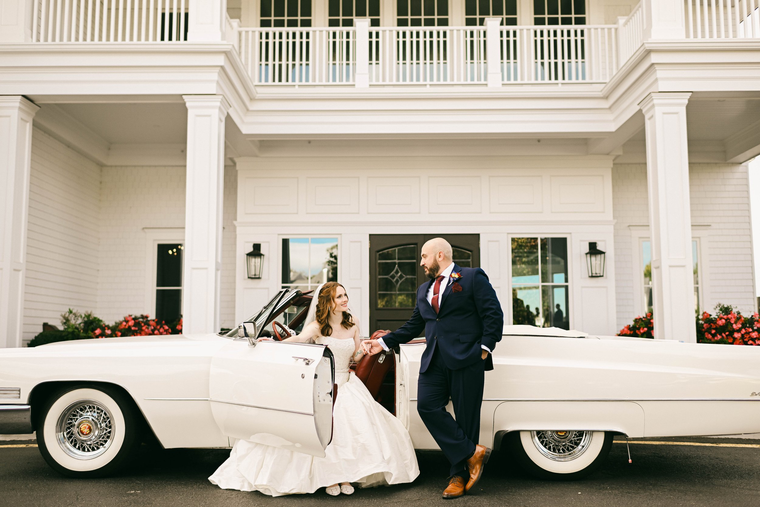A bride and groom pose in front of a white vintage convertible car parked outside a large white house with columns. The bride sits inside the car, and the groom stands outside, holding her hand.