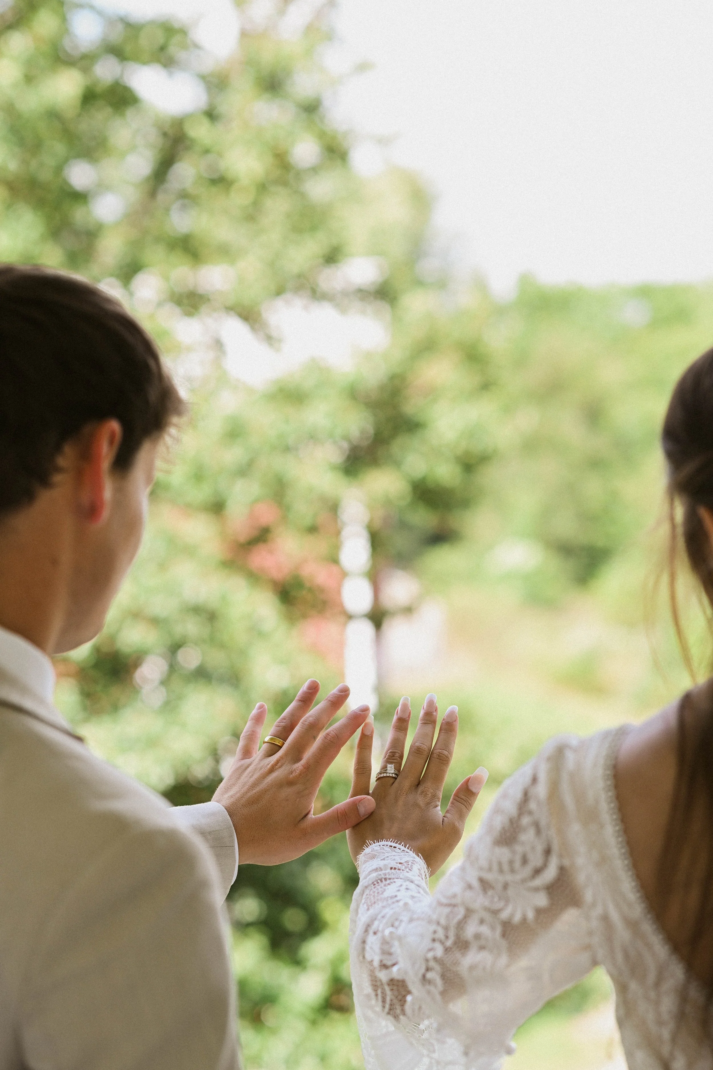 A man and a woman are outdoors, facing each other, holding hands and touching each other's fingers, with a blurred background of green trees.