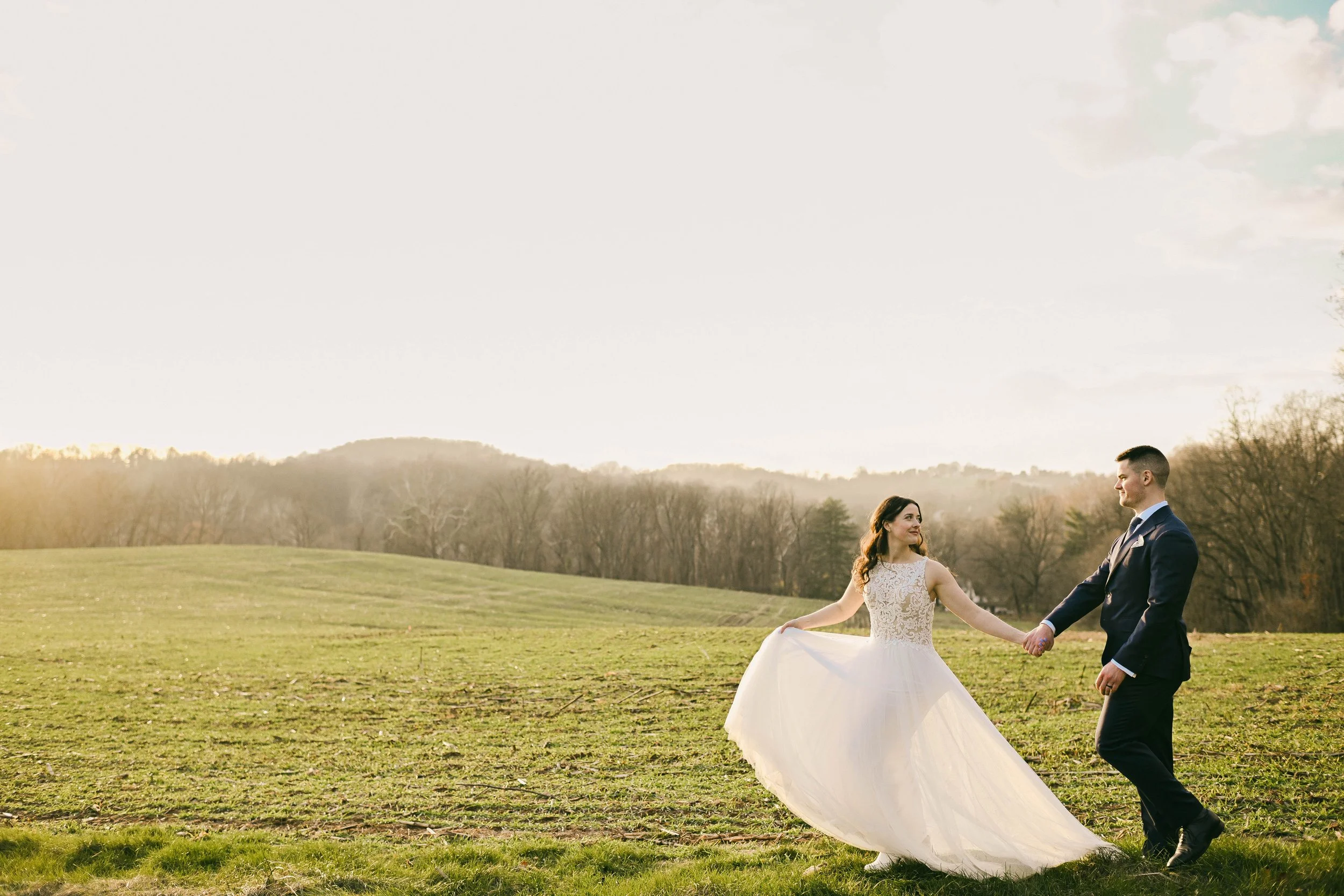 A bride and groom holding hands and dancing outdoors in a grassy field with trees and hills in the background during sunset.