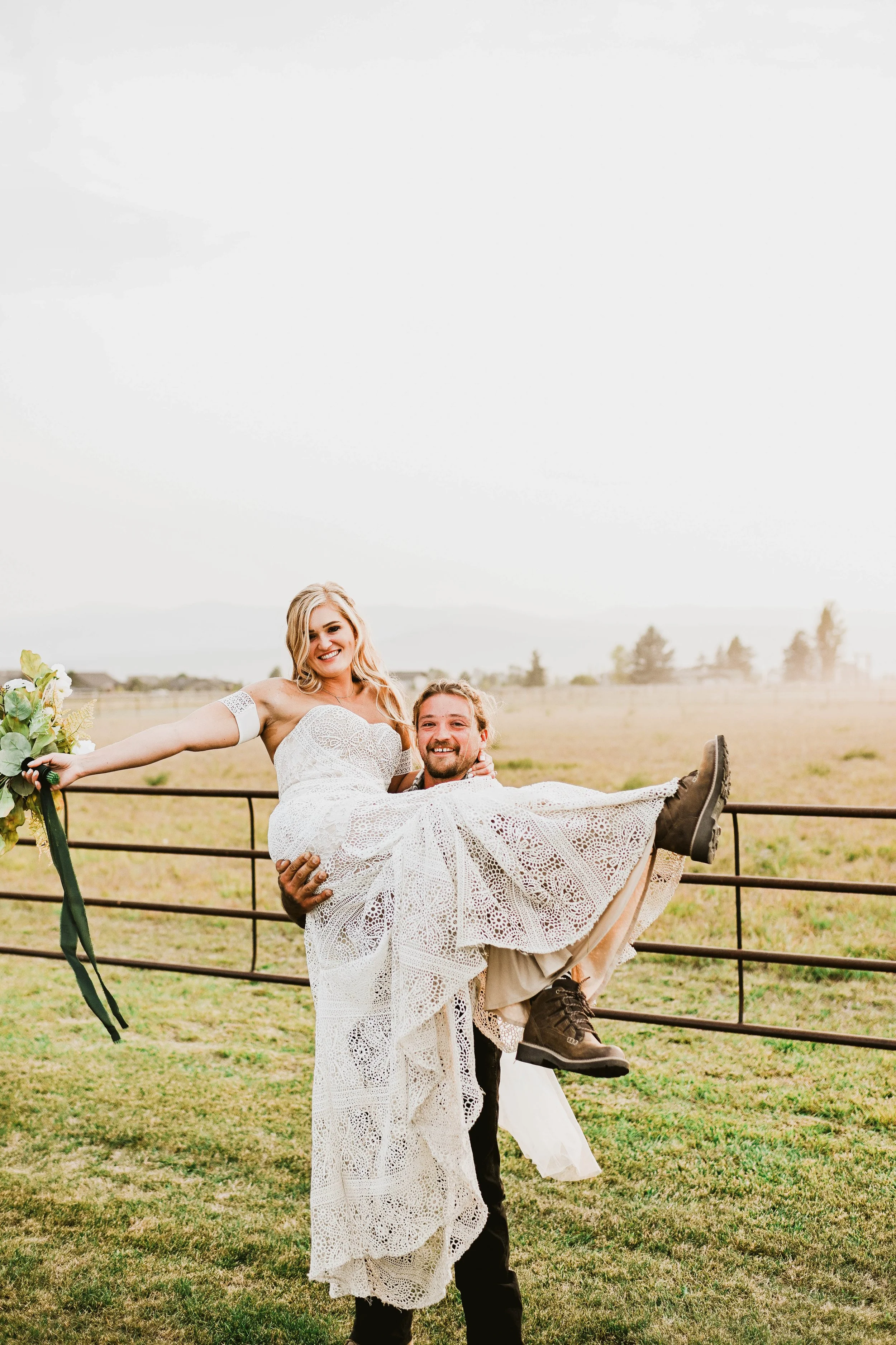 A happy couple outdoors, the man lifting the woman in a white lace dress, with a rural field and fence in the background.
