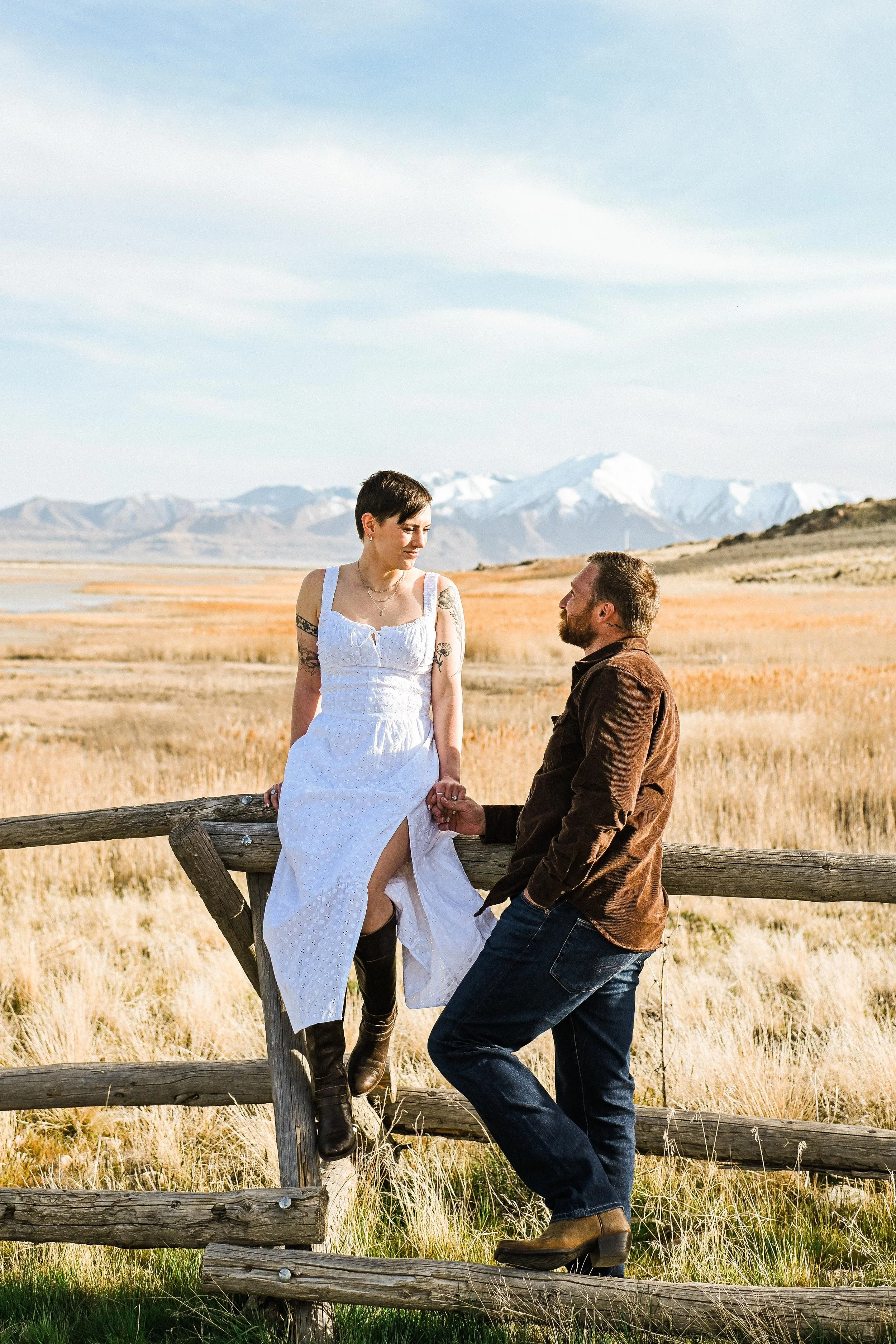 A man kneeling while holding a woman's hand in a scenic field with mountains in the background, during golden hour.