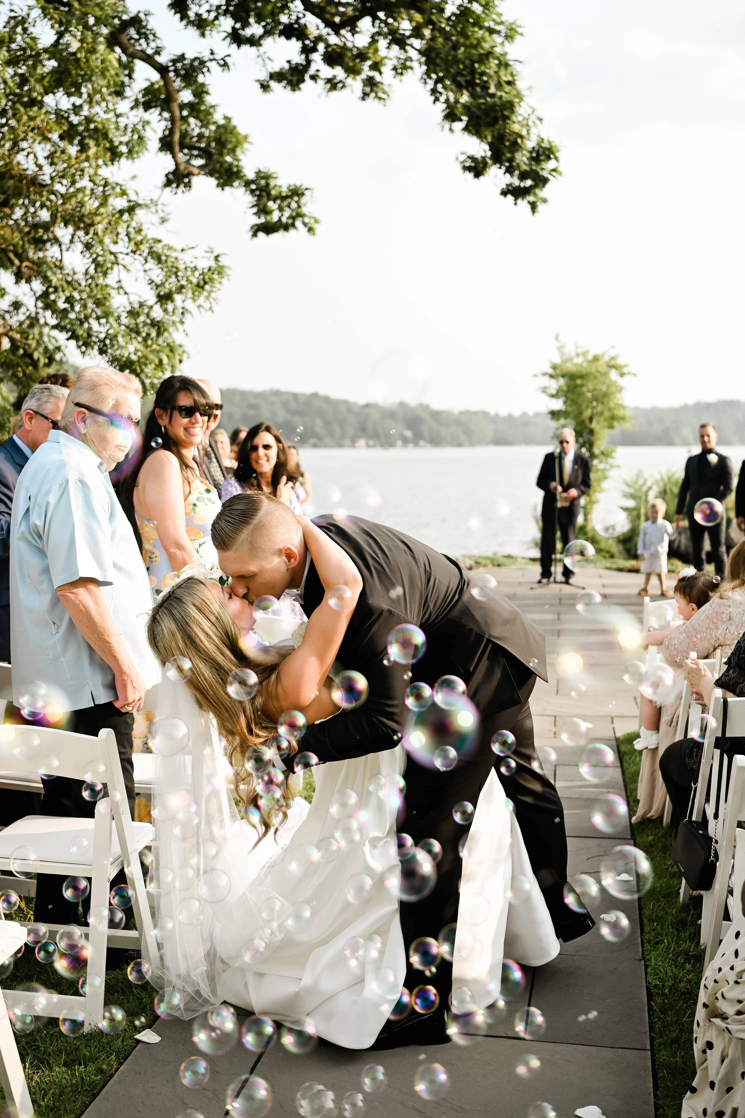 A wedding ceremony outdoors by a lake with a couple sharing a kiss, surrounded by family and guests, with bubbles floating in the air.