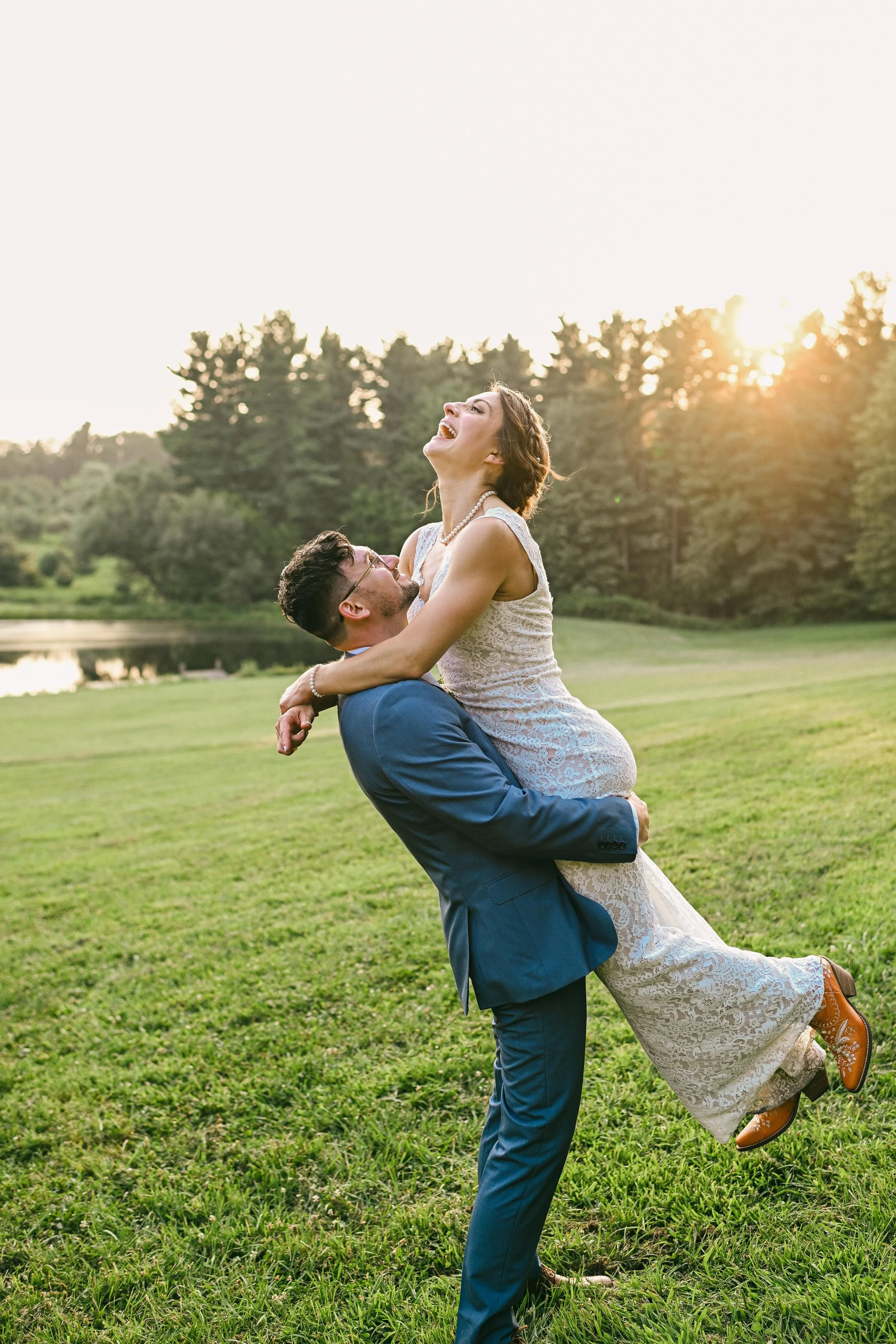 A happy couple outdoors at sunset, the man in a blue suit lifting the woman in a white lace dress, with trees and a lake in the background.