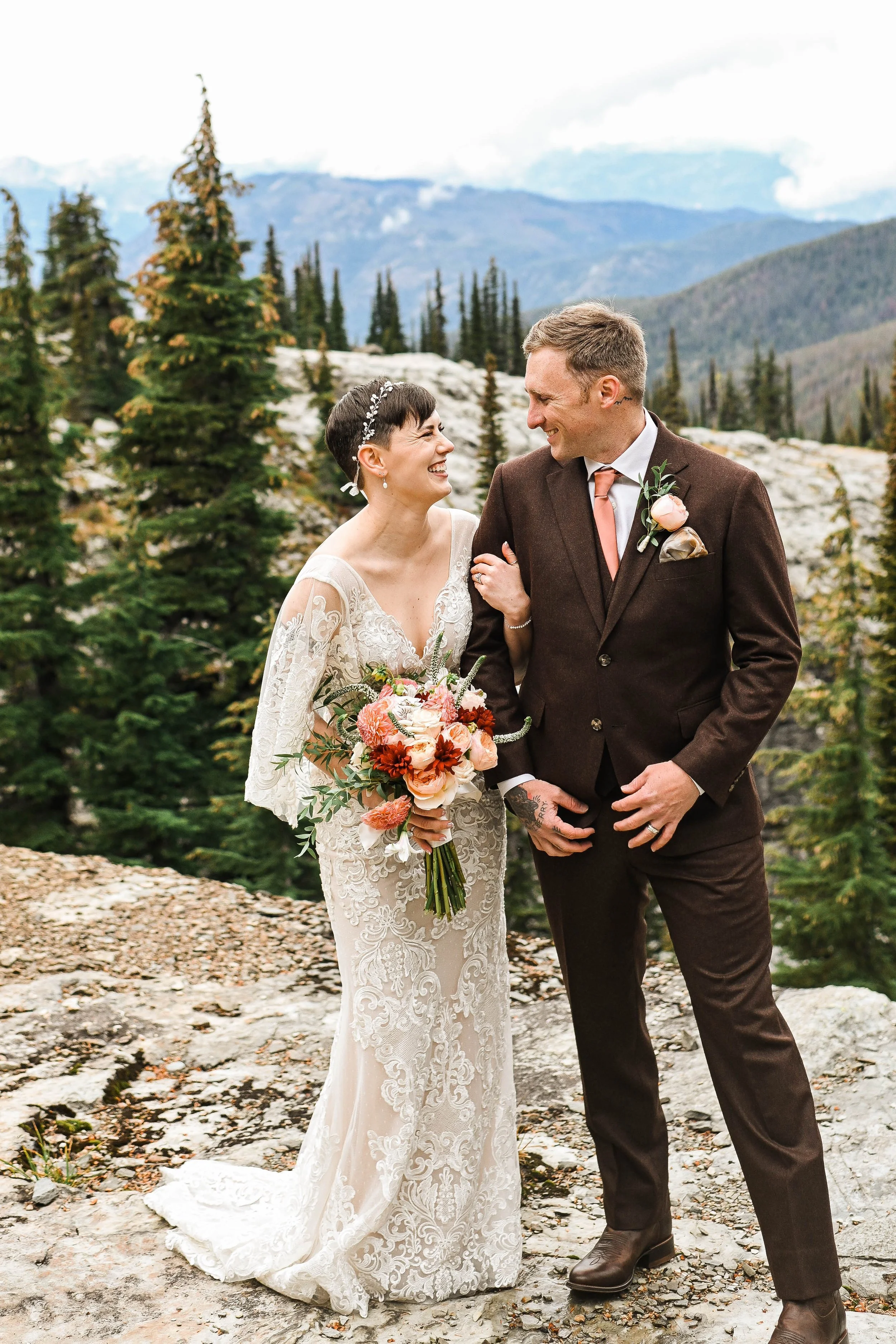 A bride and groom in wedding attire smiling and looking at each other outdoors with a mountainous landscape and trees in the background.
