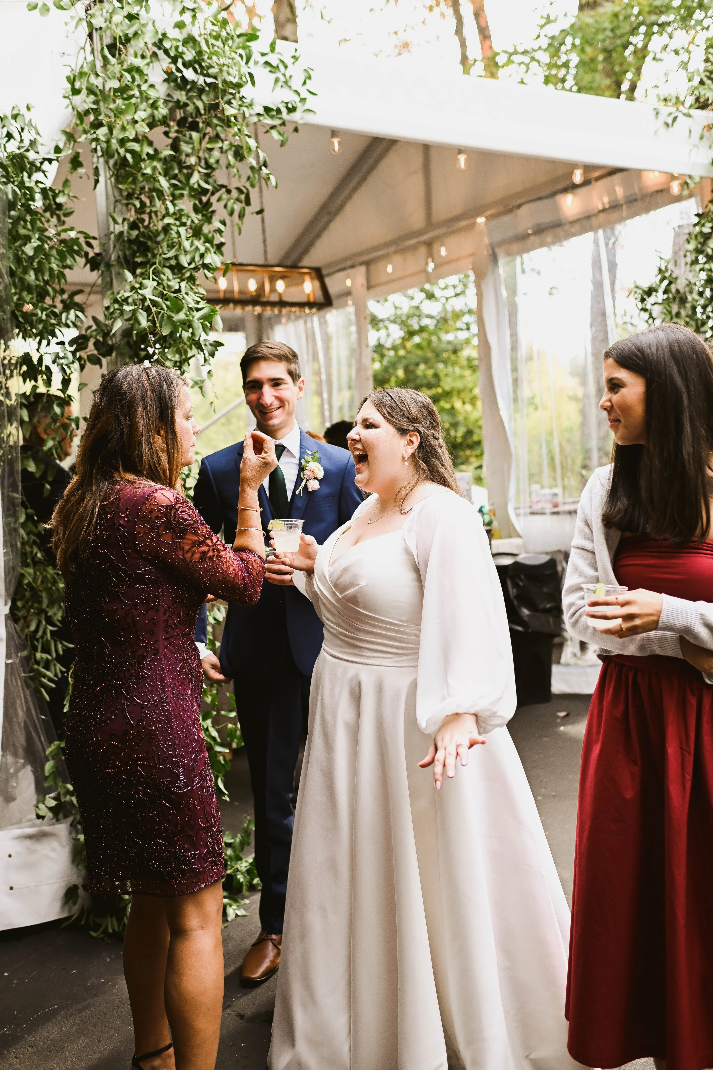 People celebrating at a wedding reception, with a woman in a white dress laughing and talking to a woman in a burgundy dress, a man in a blue suit, and another woman in a red dress holding drinks, under a canopy with string lights and greenery.