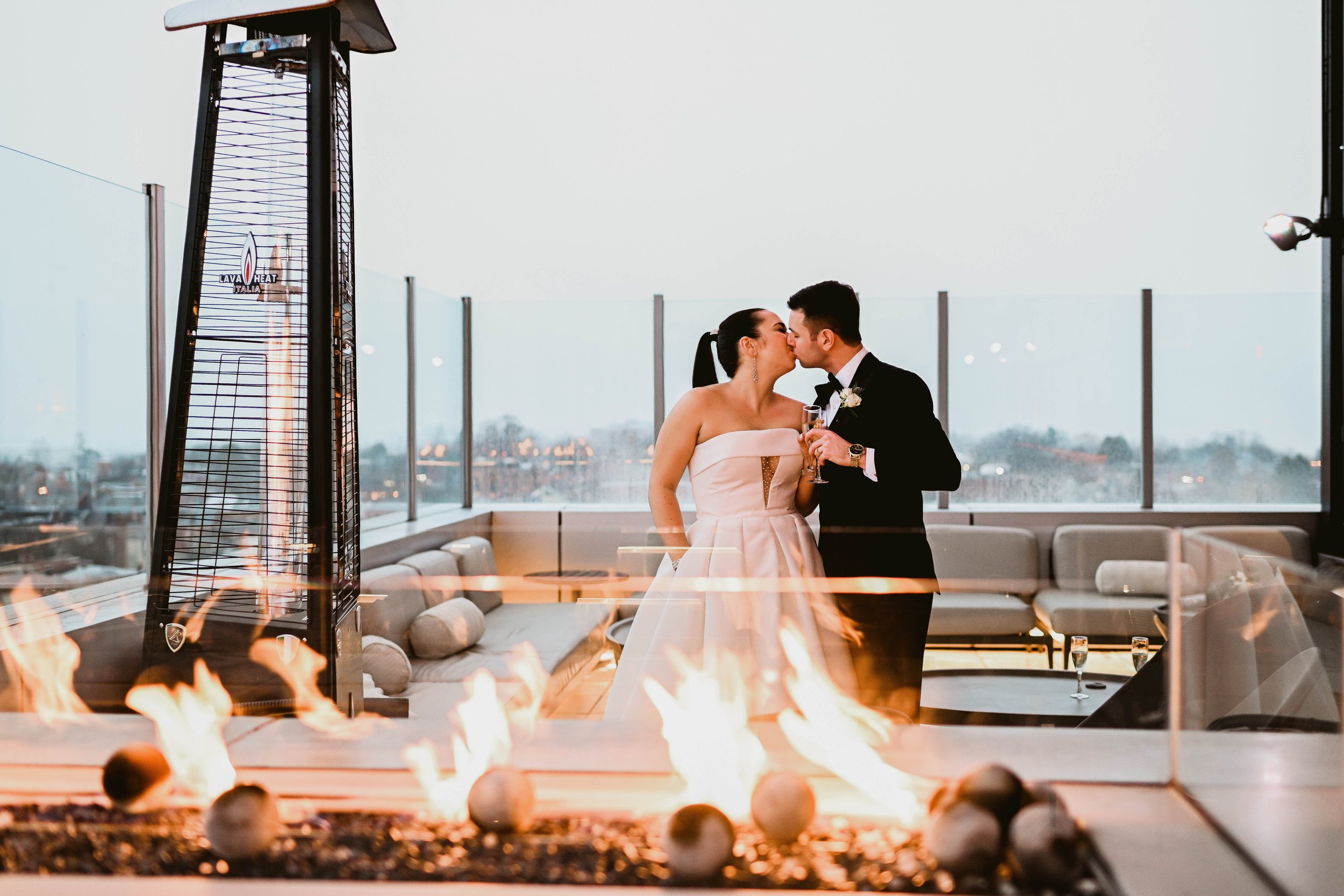 A newlywed couple sharing a kiss at their wedding reception, standing next to a fireplace, with cityscape view through large windows in the background.