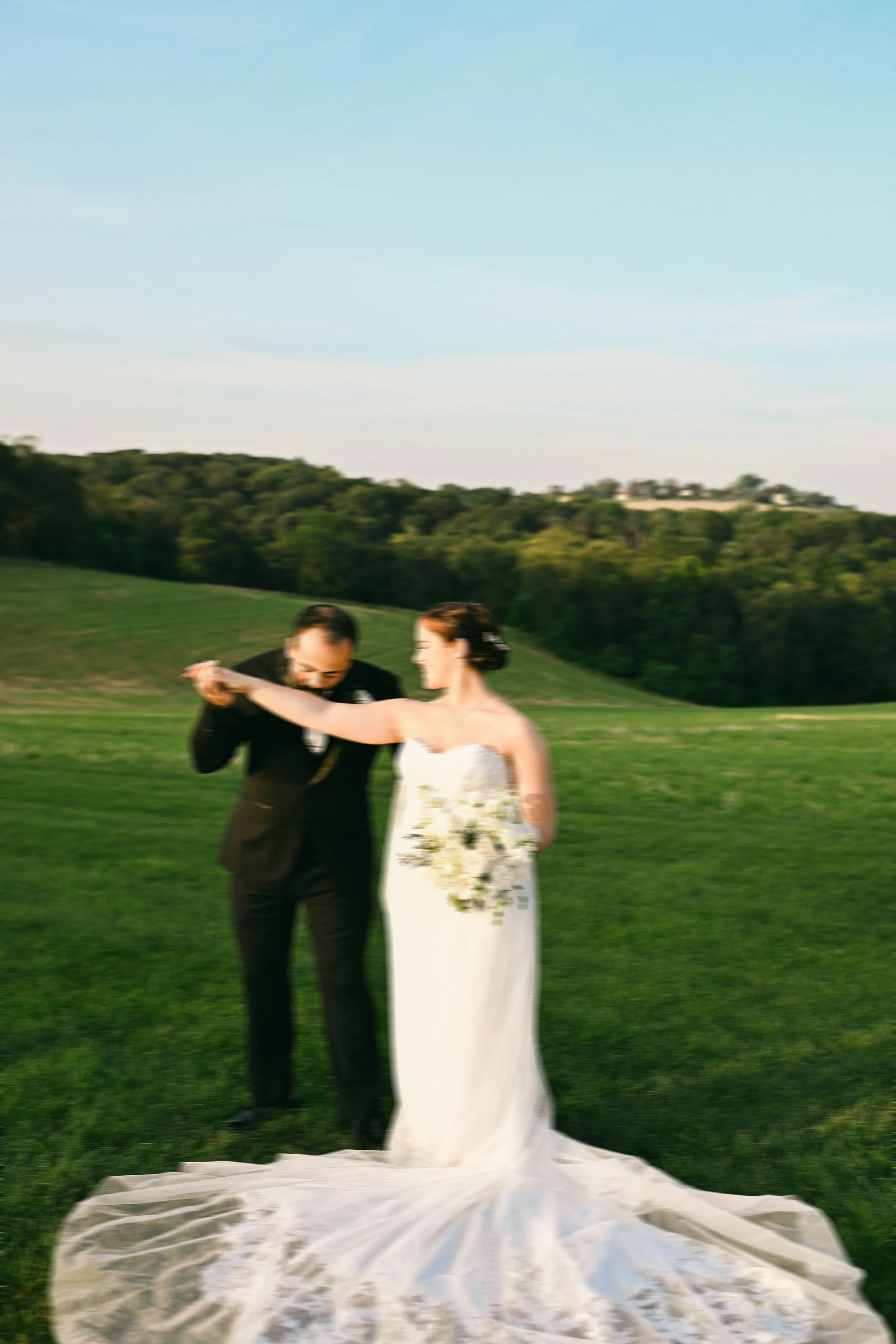 A bride and groom dancing outdoors on a grassy field during sunset, with trees and hills in the background.