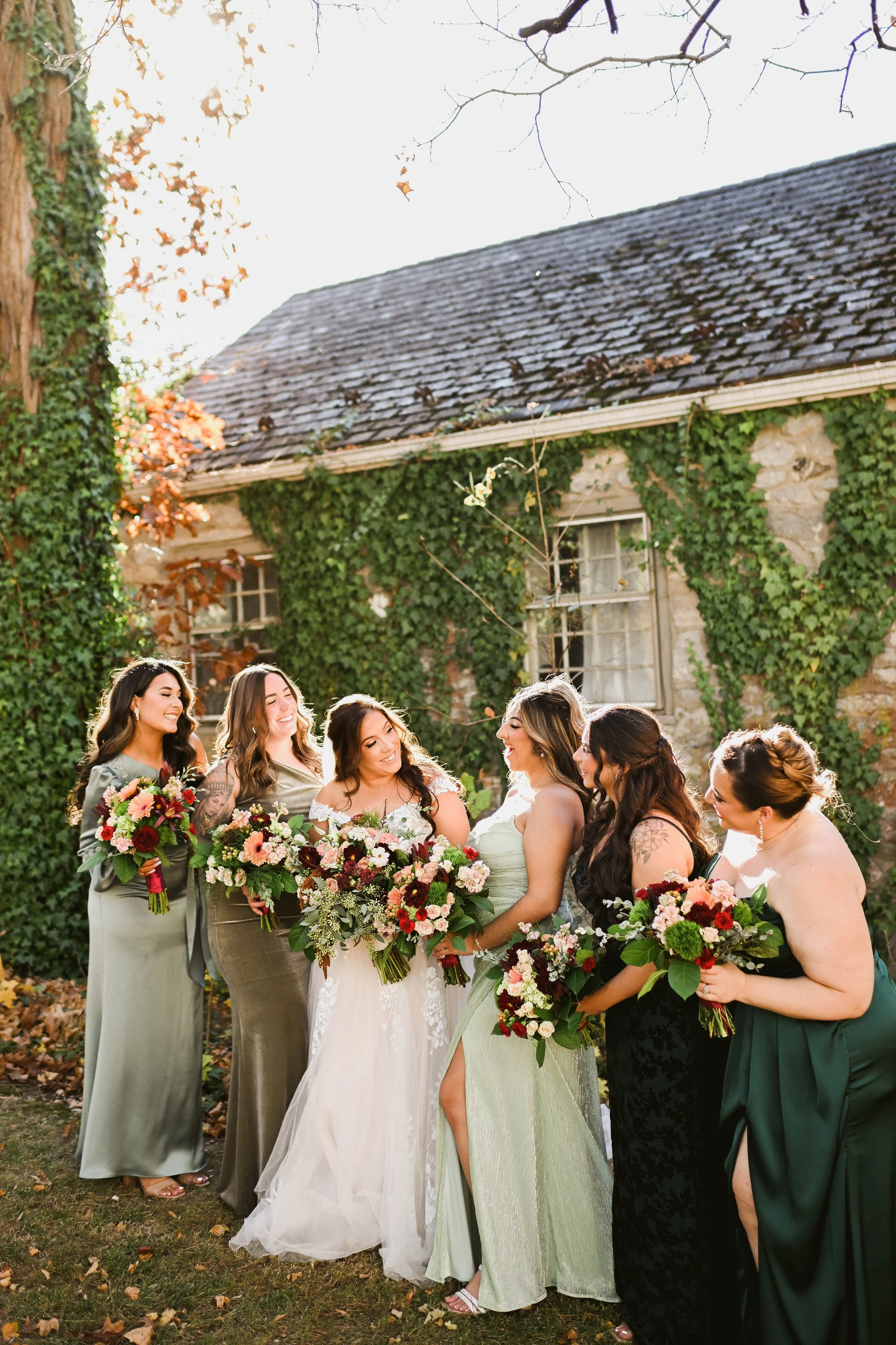 A group of six women in dresses holding bouquets of flowers, smiling and standing outdoors in front of a stone house covered in ivy, during daytime.