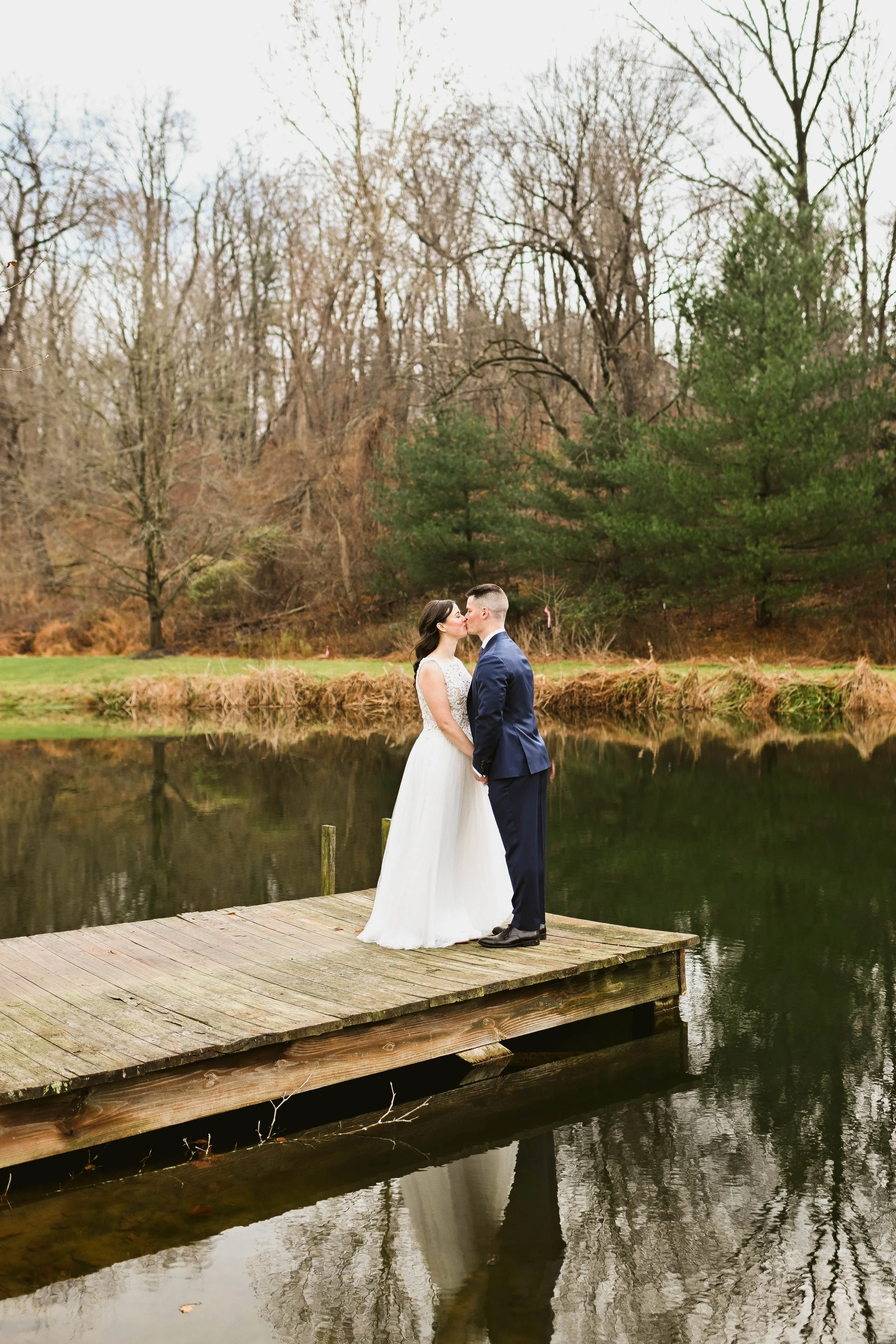 A bride and groom kiss on a wooden dock by a pond, with trees in the background on a cloudy day.