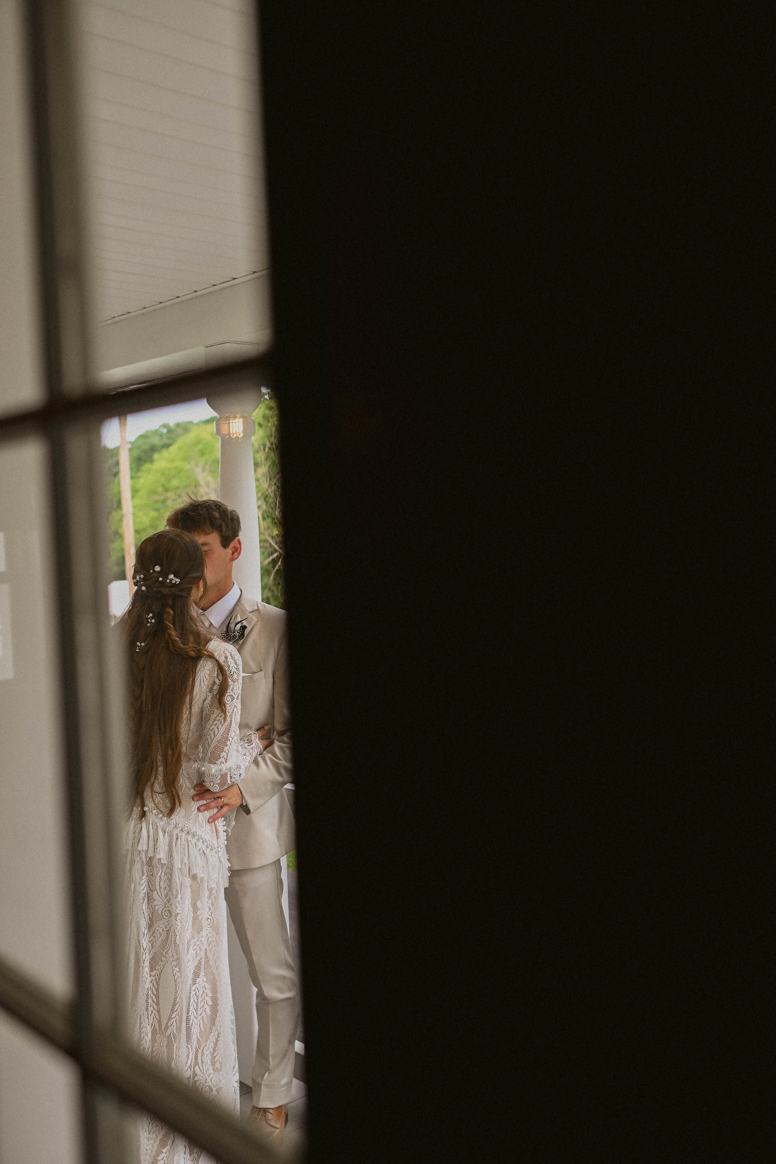 A couple dressed in wedding attire stands closely outside under a covered porch, sharing an intimate moment, seen through a narrow gap between part of a building and a dark panel.