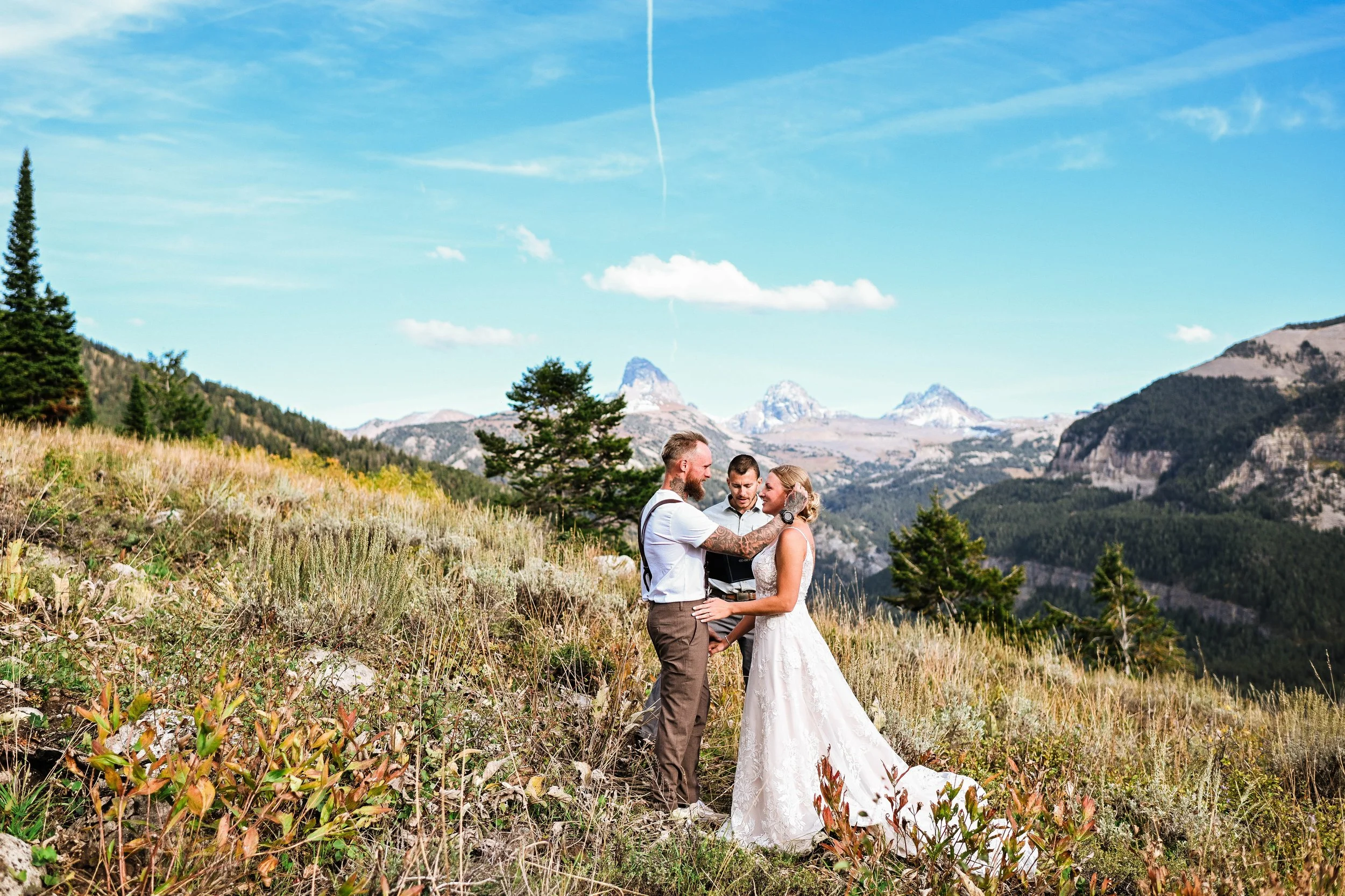 A wedding couple standing outdoors on a grassy hillside with mountains in the background, exchanging vows while an officiant stands between them under a blue sky.