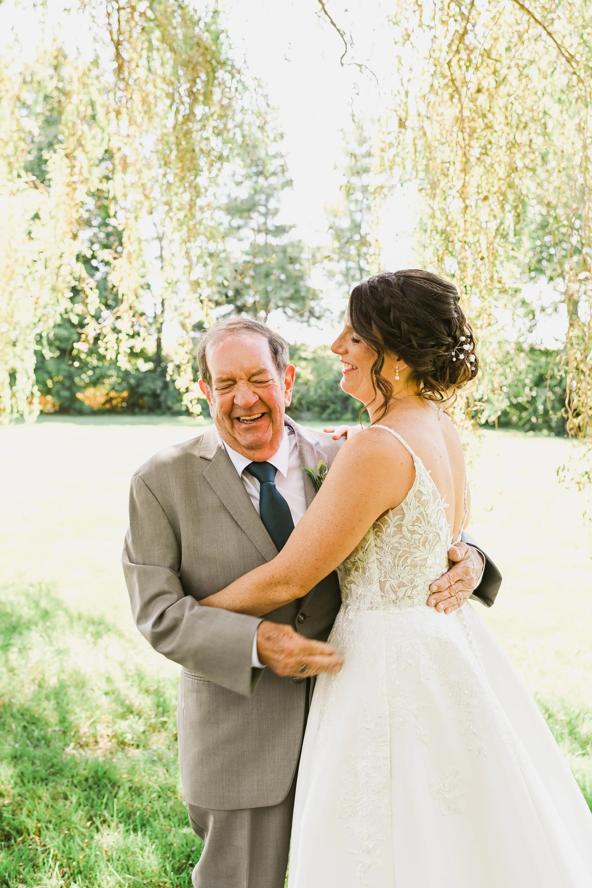 A bride and an older man are smiling and dancing outdoors in a park with green trees and grass, under a hanging branch.