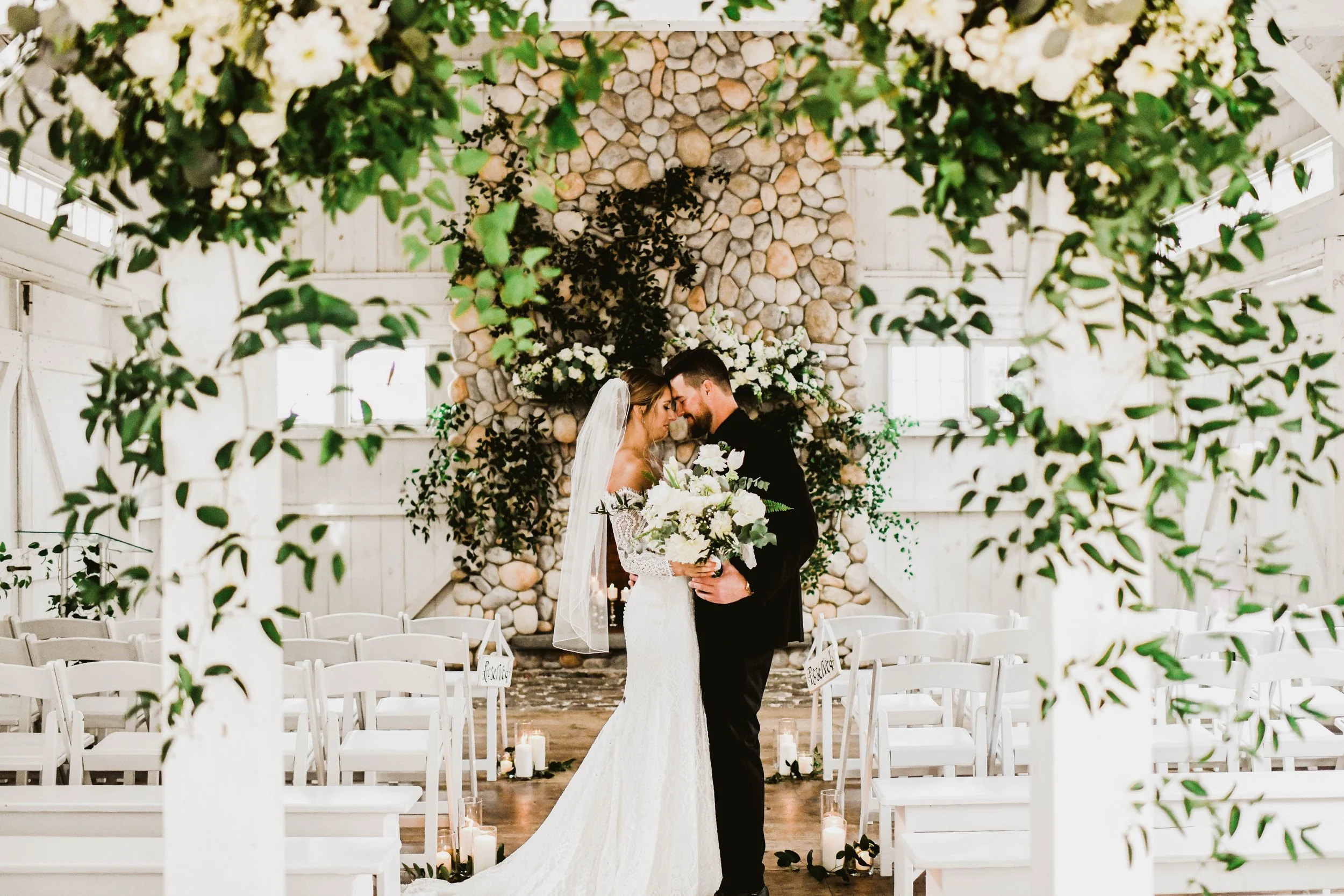 A bride and groom standing close together with foreheads touching inside a decorated wedding venue. The bride is holding a bouquet of white flowers and wearing a white lace wedding dress and veil, while the groom is in a black suit. The background fe