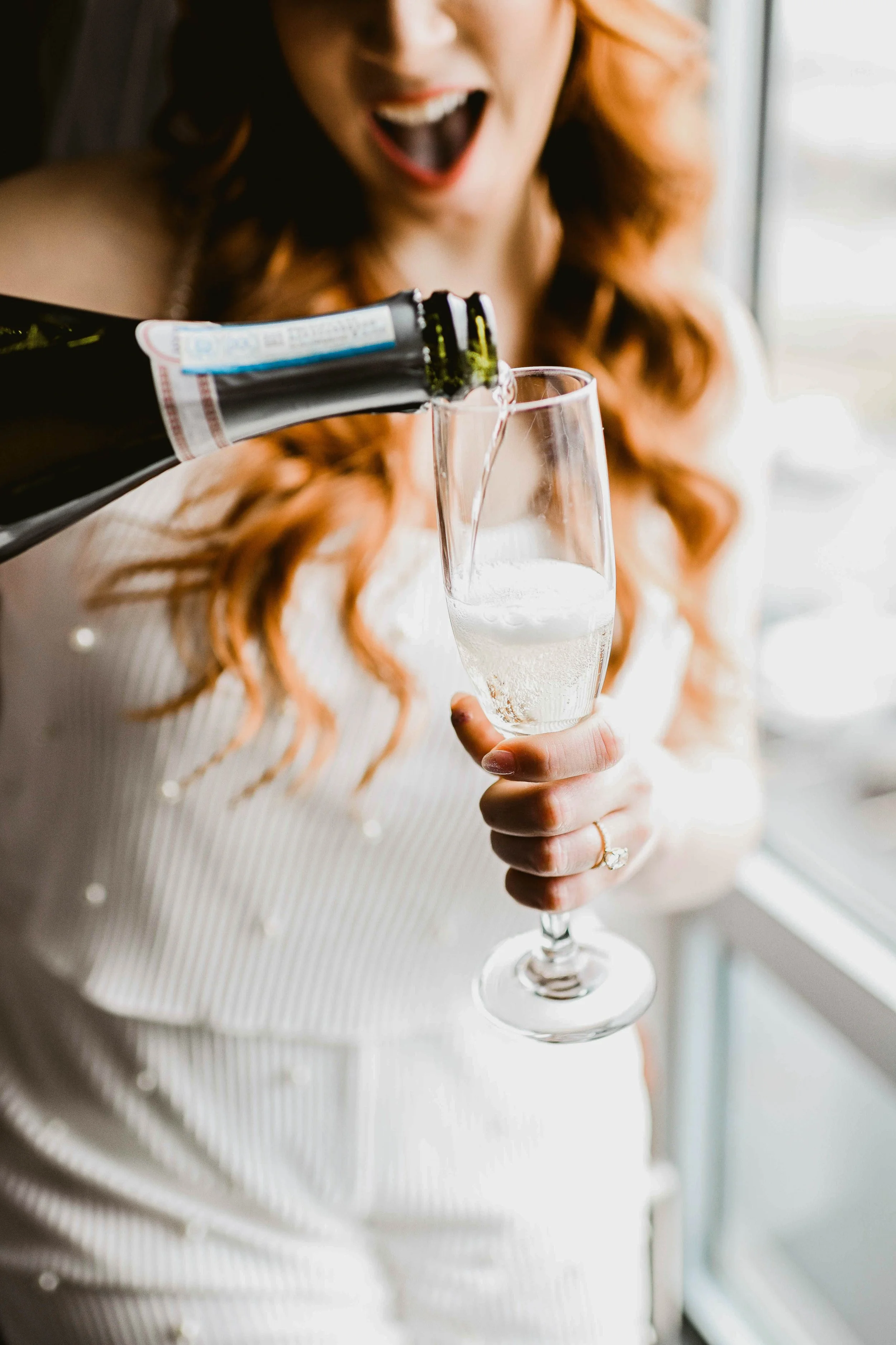 A woman with red hair in a white outfit is pouring champagne into a flute.