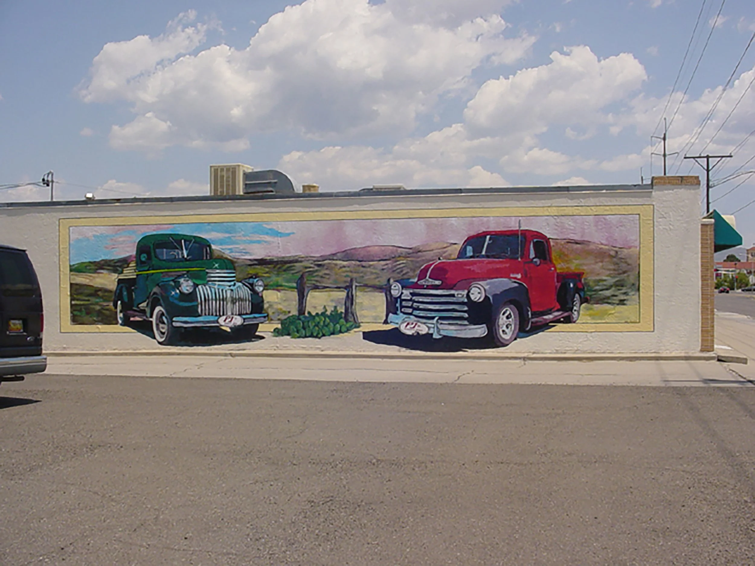 Colorful mural of two vintage pickup trucks, one green and one red, painted on a building wall with a landscape background and a blue sky with clouds.
