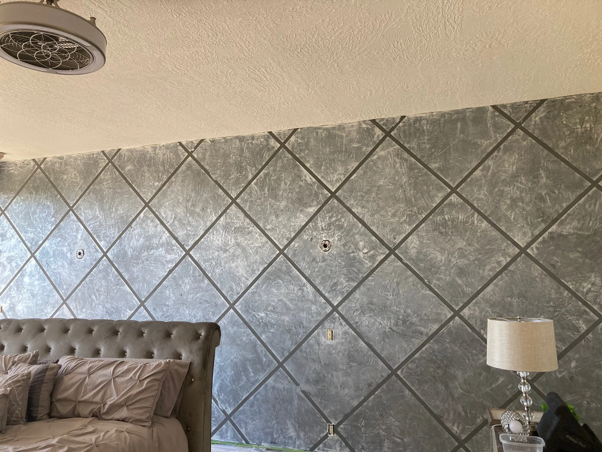 A bedroom wall decorated with dark grey textured tiles arranged in a diamond pattern. Part of a bed with beige tufted headboard and beige bedding is visible in the lower left. There is a nightstand with a beige lamp, a black item, and some decorative