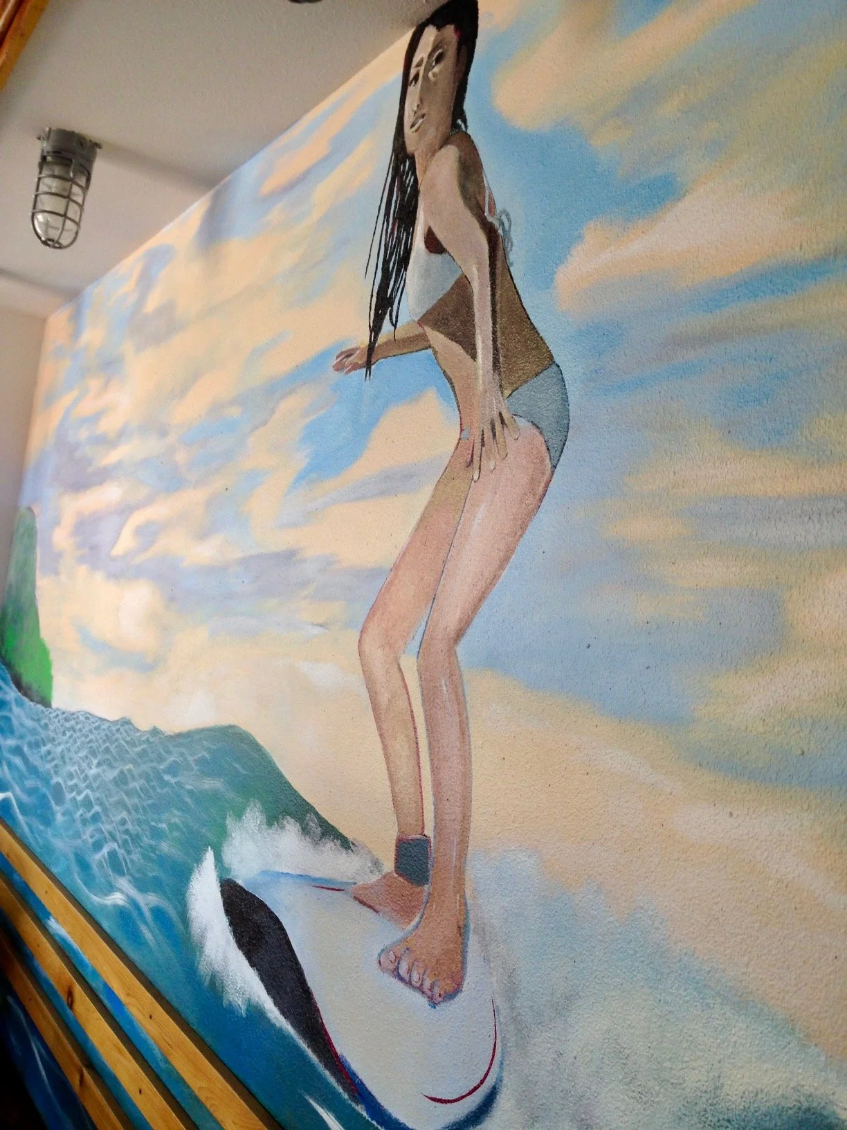A mural of a woman in a bikini riding a surfboard on a wave with a sky background.