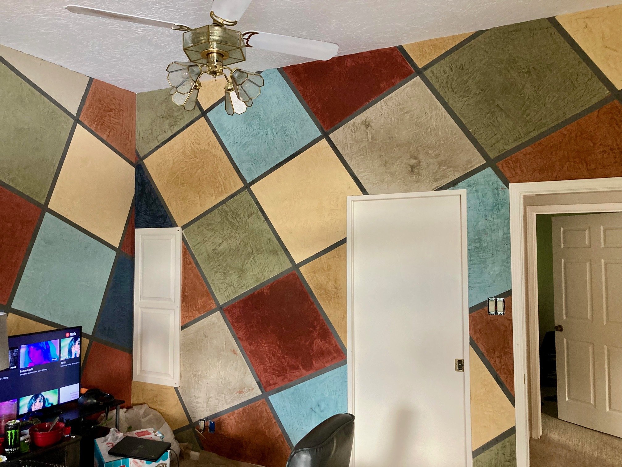 Room with colorful geometric painted walls, flat ceiling, ceiling fan, white doors, and a television showing music videos.
