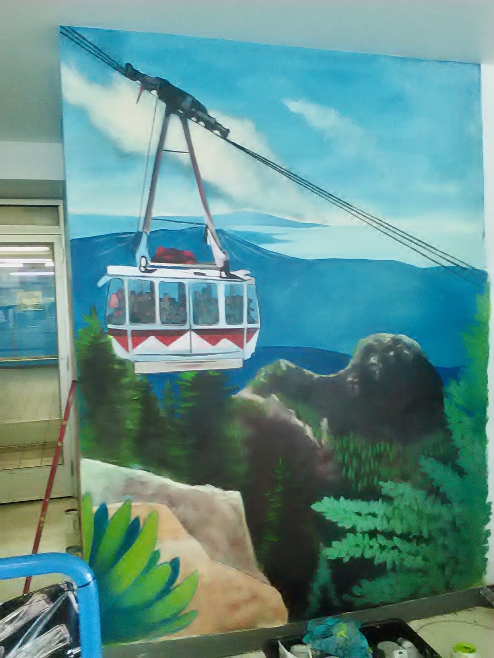 Colorful mural of a cable car traveling above a mountainous forested landscape with trees, rocks, and a blue sky.
