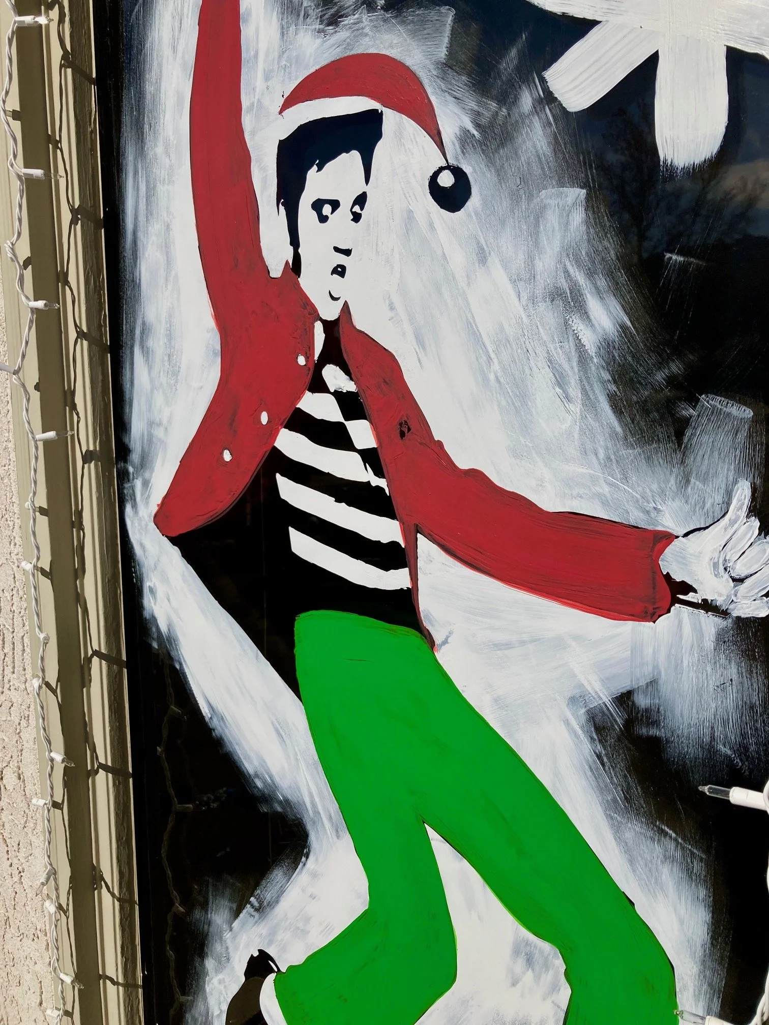 Stylized painting of a young man who looks like Elvis Presley with a Christmas Santa Hat, wearing a red jacket, black and white striped shirt, and bright green pants, set against a dynamic black and white background.  Billings, Montana custom murals.